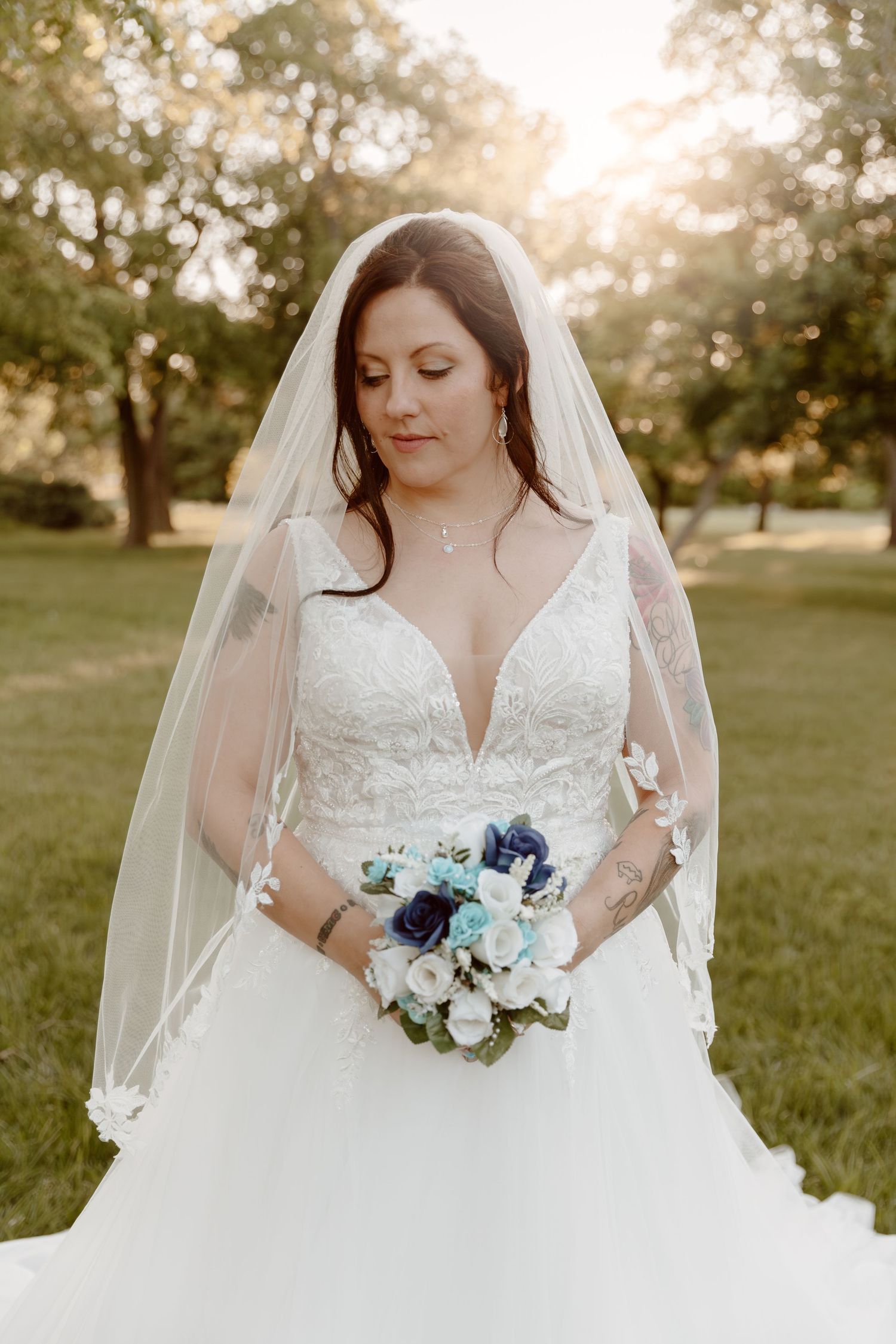A bride in a flowing white lace gown holds a blue and white bouquet during golden hour outdoor bridal portraits.