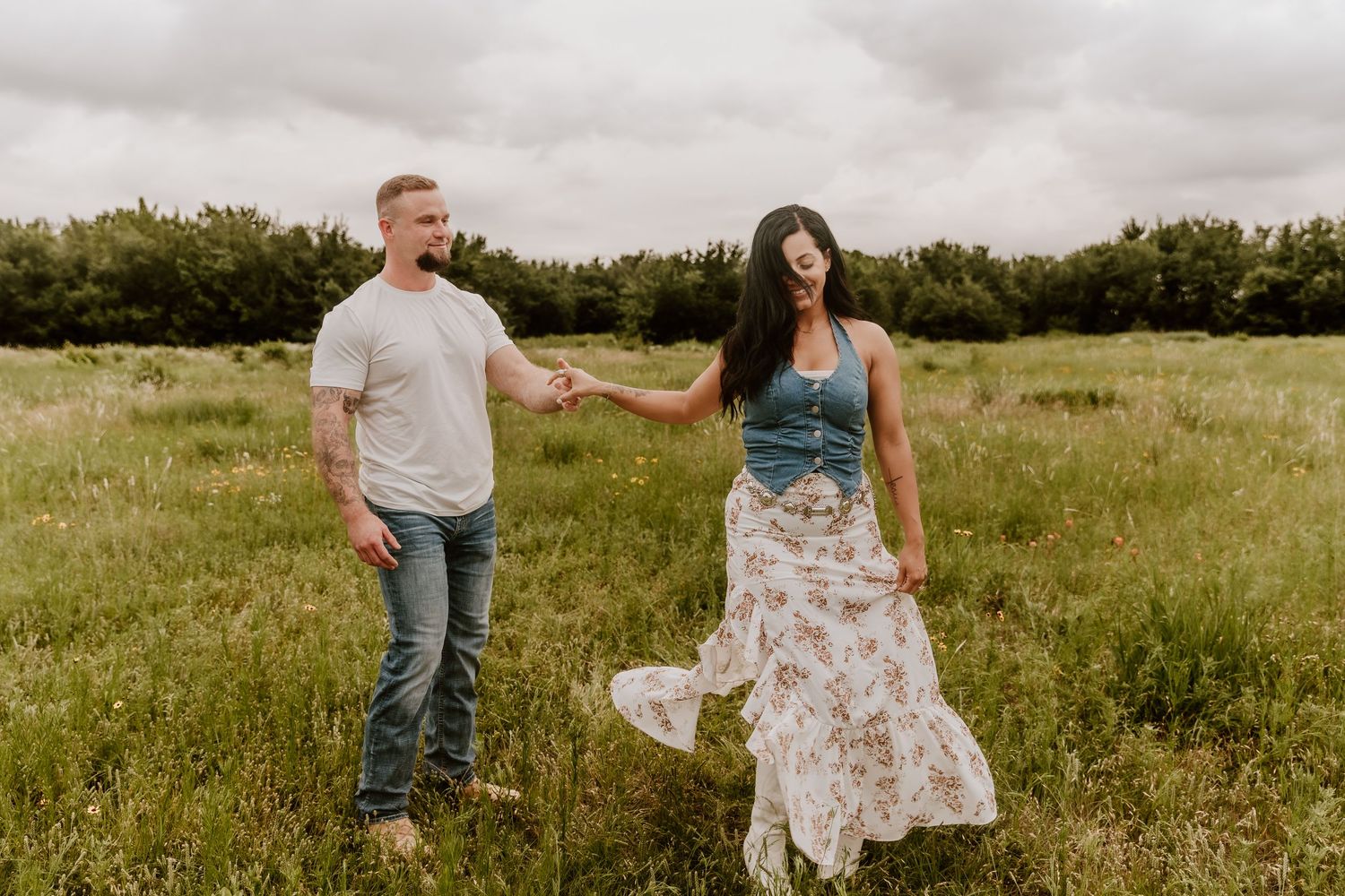 Couple holding hands walking through a lush green meadow under cloudy skies, woman in floral skirt twirling.