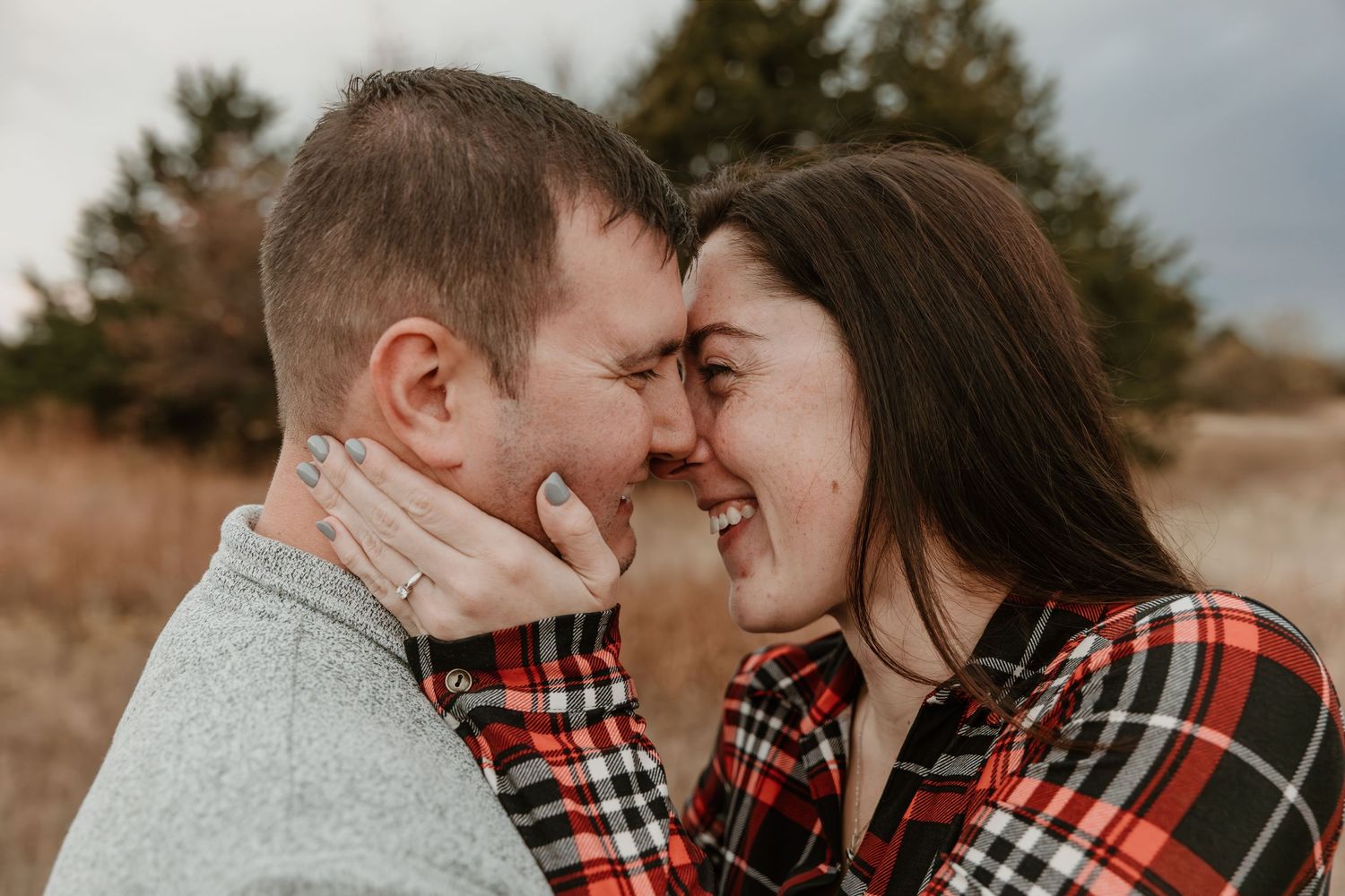A couple wearing coordinated red plaid and grey outfits embrace in a golden autumn field during sunset.