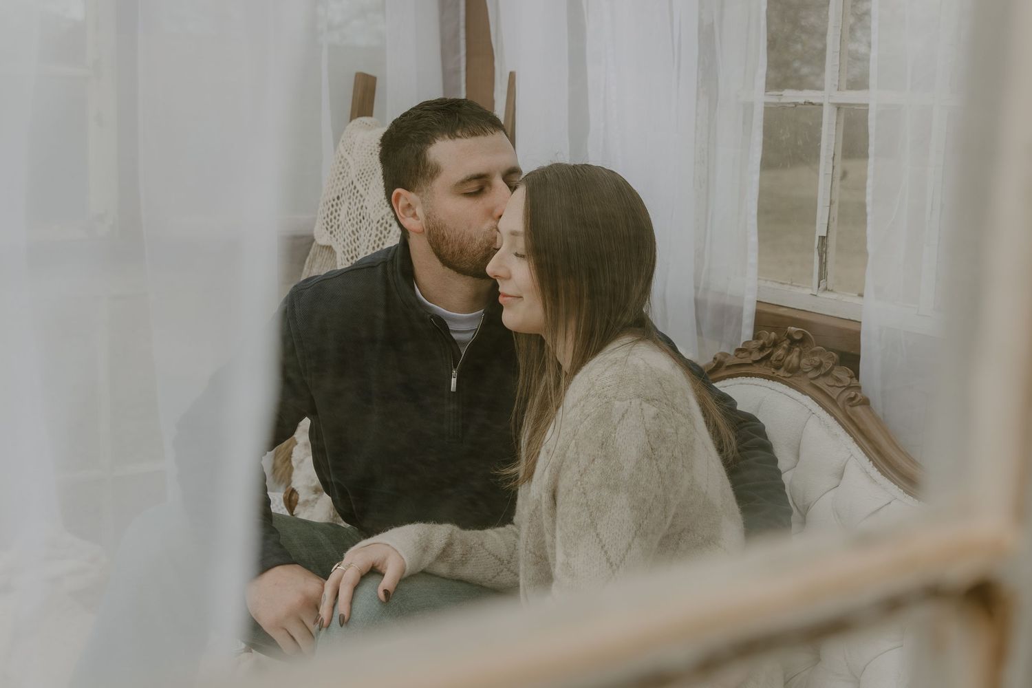 A couple sits close together on a couch in a bright living room during an intimate moment.