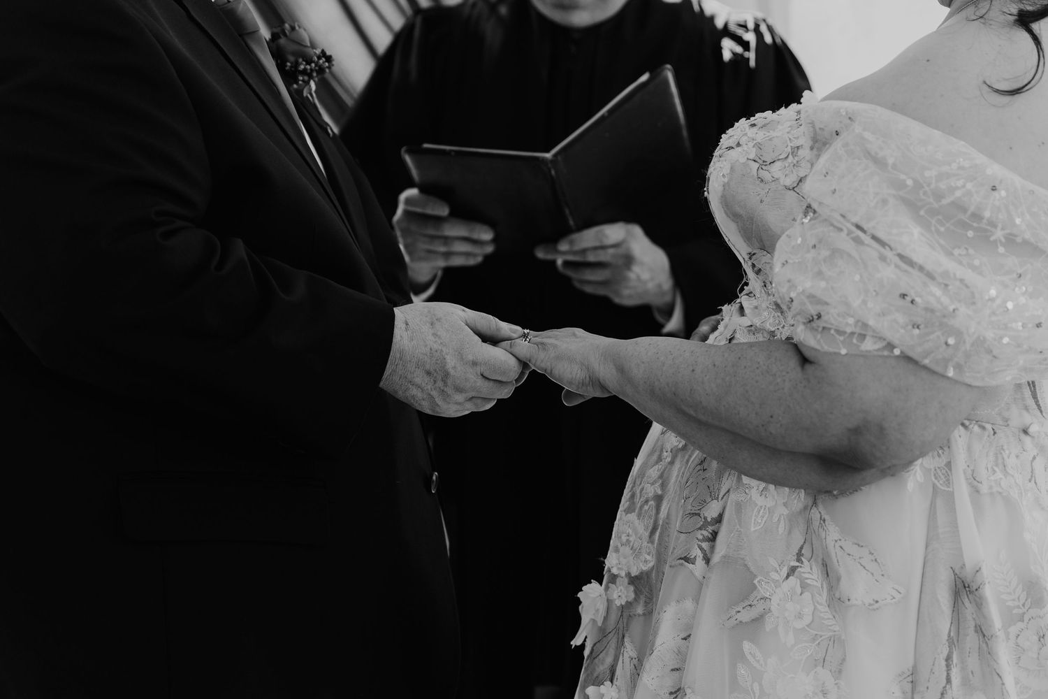 Black and white wedding ceremony photo showing hands exchanging rings during traditional vow reading.