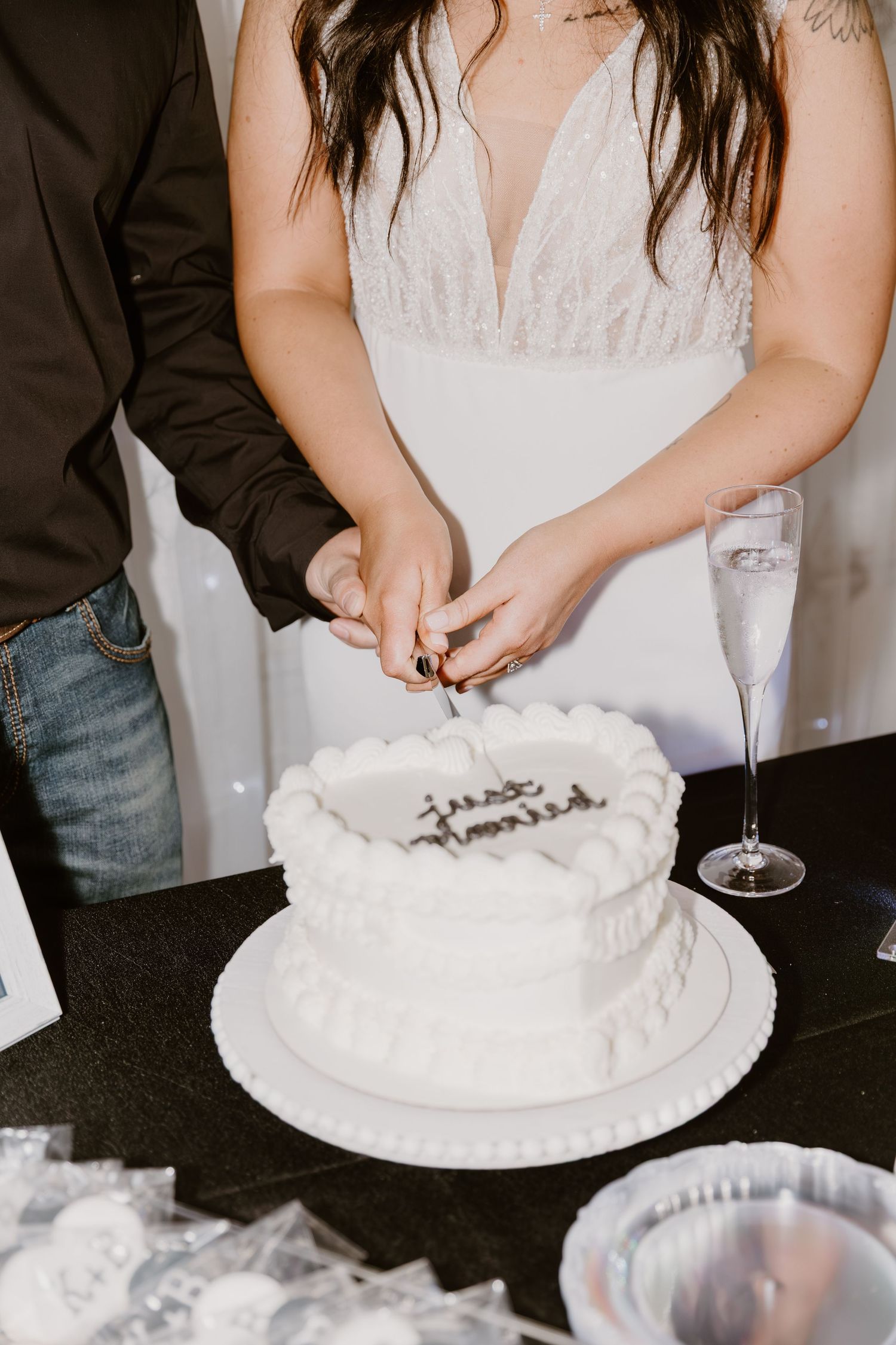 A couple cutting their white wedding cake together while holding champagne glasses during their reception celebration.