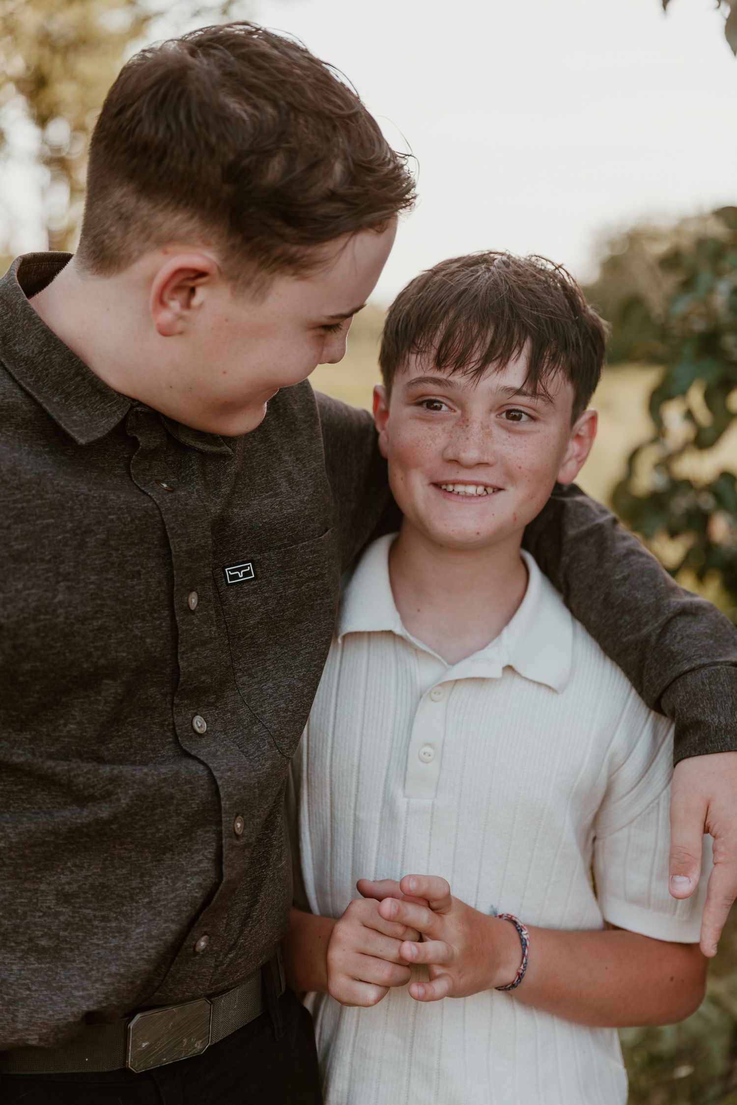 Two young boys in dark and light colored shirts share a close bonding moment outdoors in warm lighting.