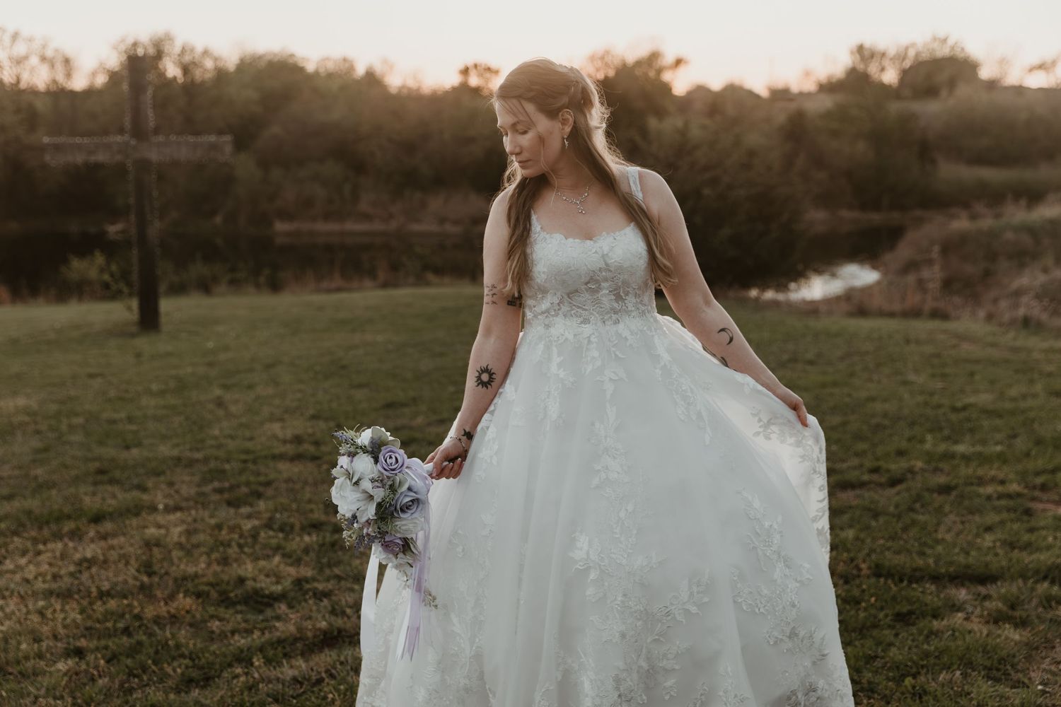Bride in strapless lace ball gown holding bouquet at golden hour outdoors near wooden cross.