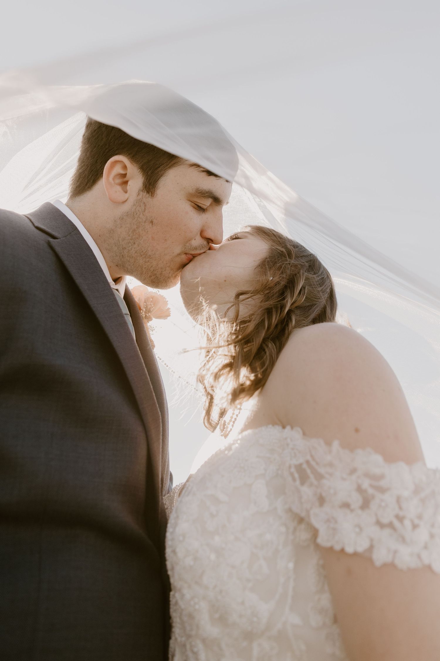 Romantic bride and groom share a kiss under flowing white veil in soft golden natural light.
