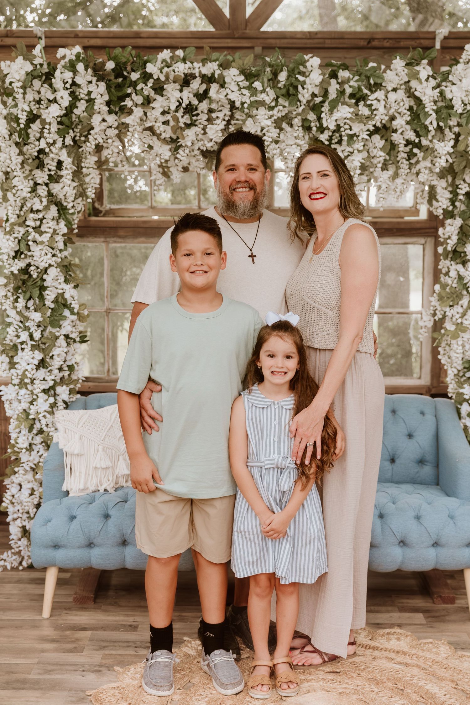 Family of four posing in front of floral arch with white blooms and blue sofa in a bright, elegant studio setting.