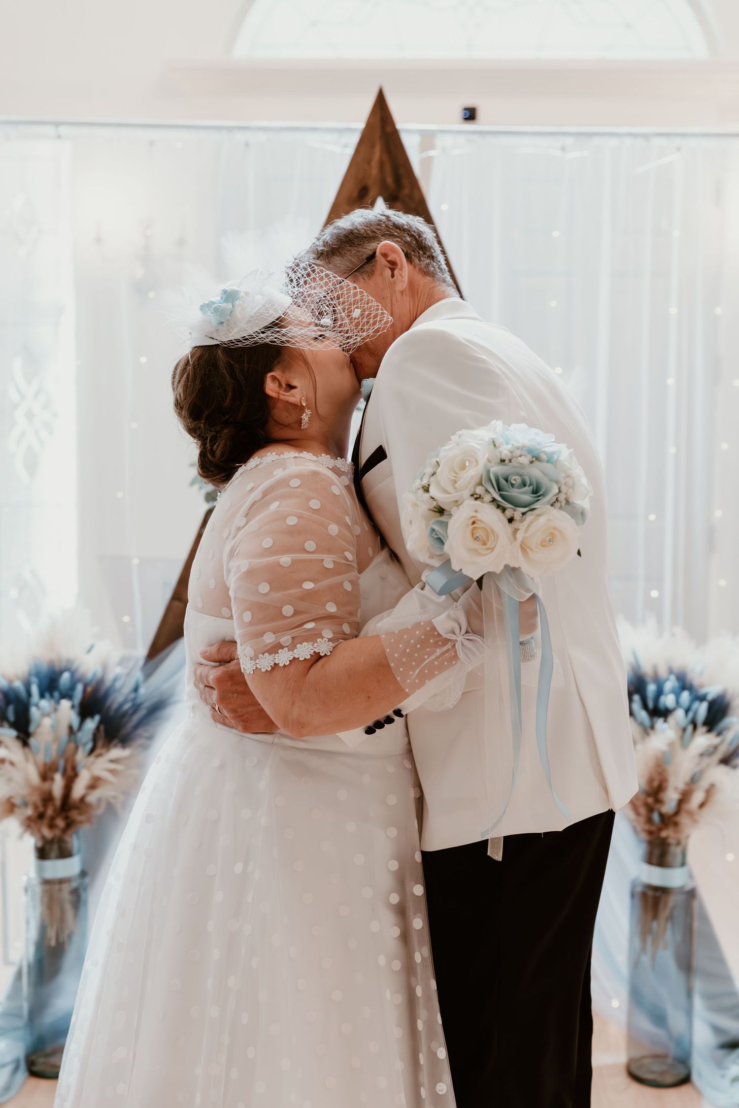 Couple sharing tender kiss during wedding ceremony with soft light and floral decor in background.