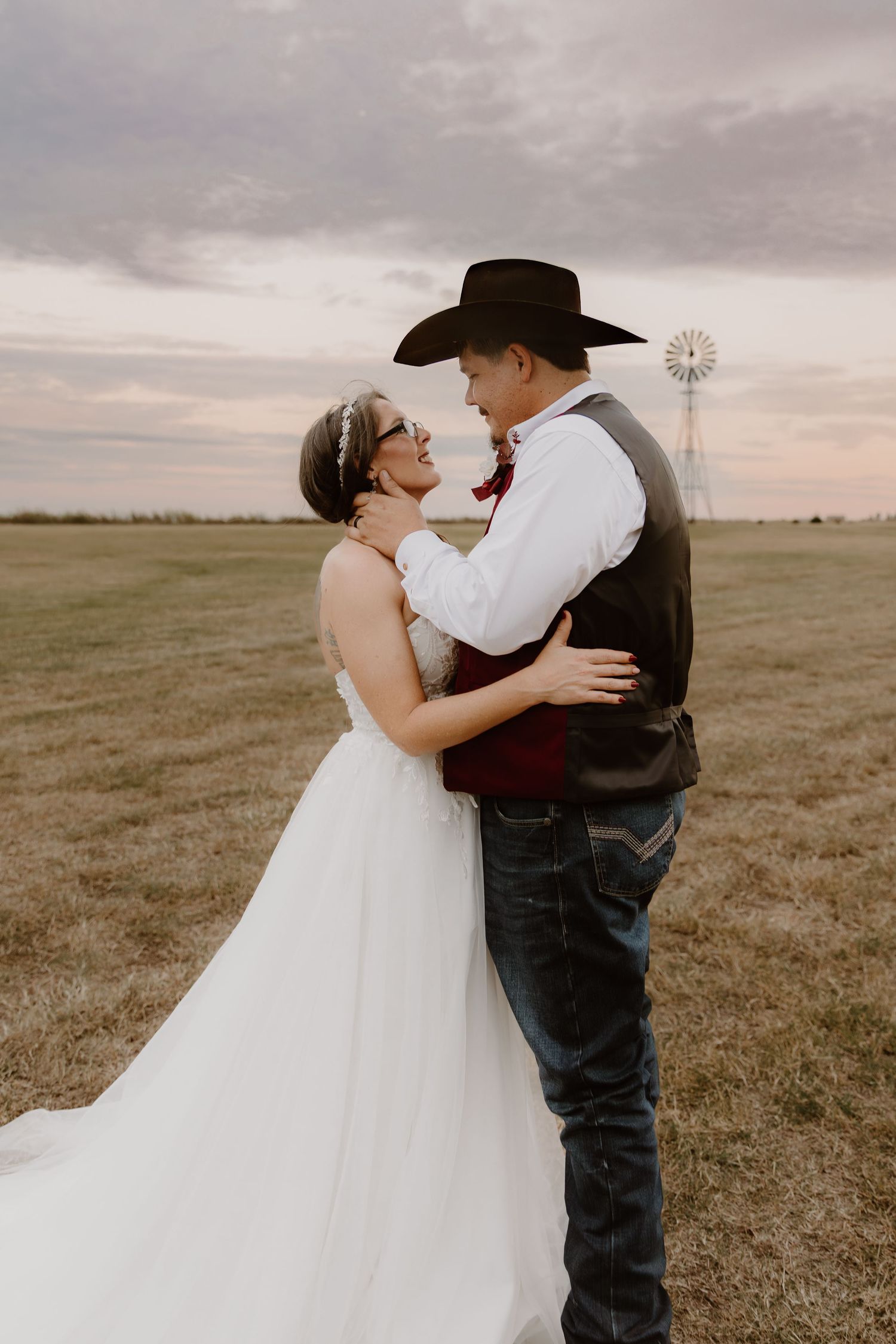 A romantic country wedding photo with a bride in white and groom in western attire embracing in a field at sunset.