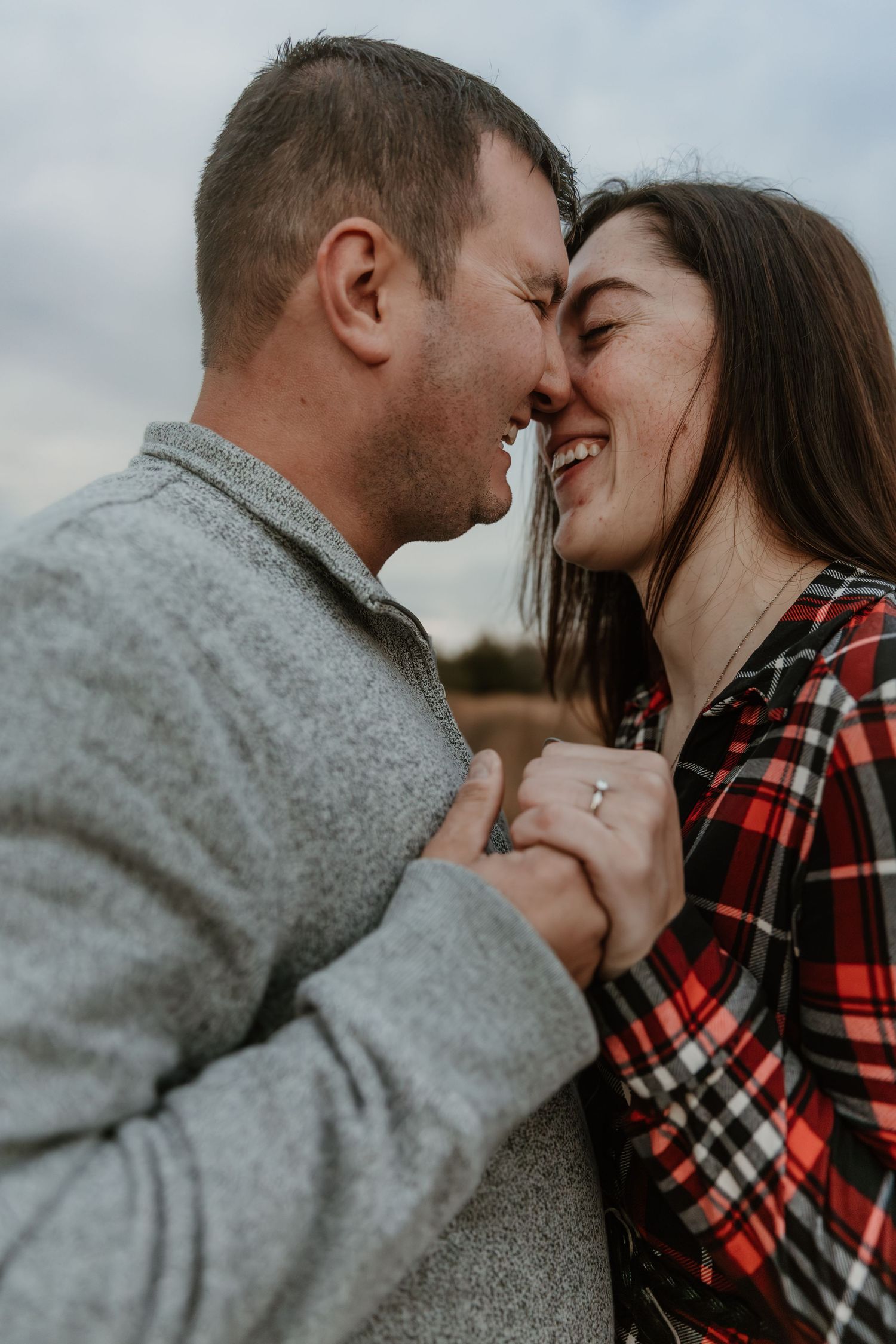 A romantic couple sharing an intimate moment outdoors, with one wearing a grey sweater and the other in a plaid shirt.