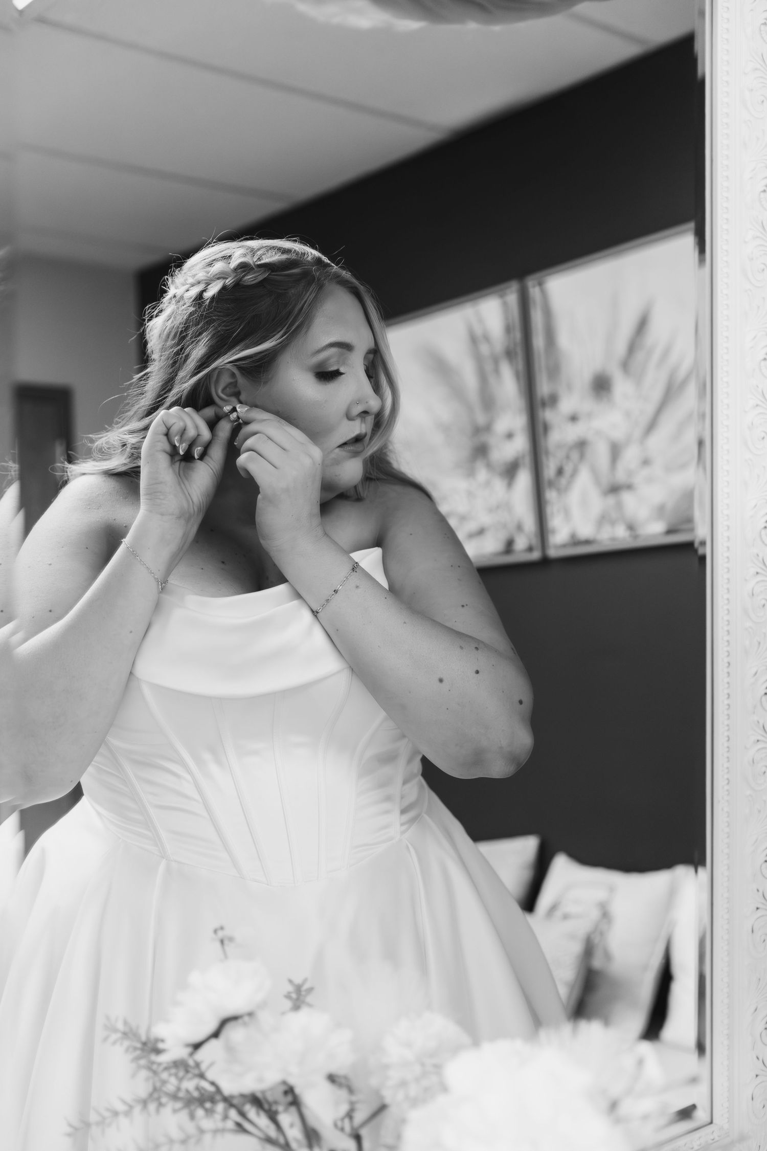 A bride in a white dress puts on her earrings while getting ready in black and white photography.