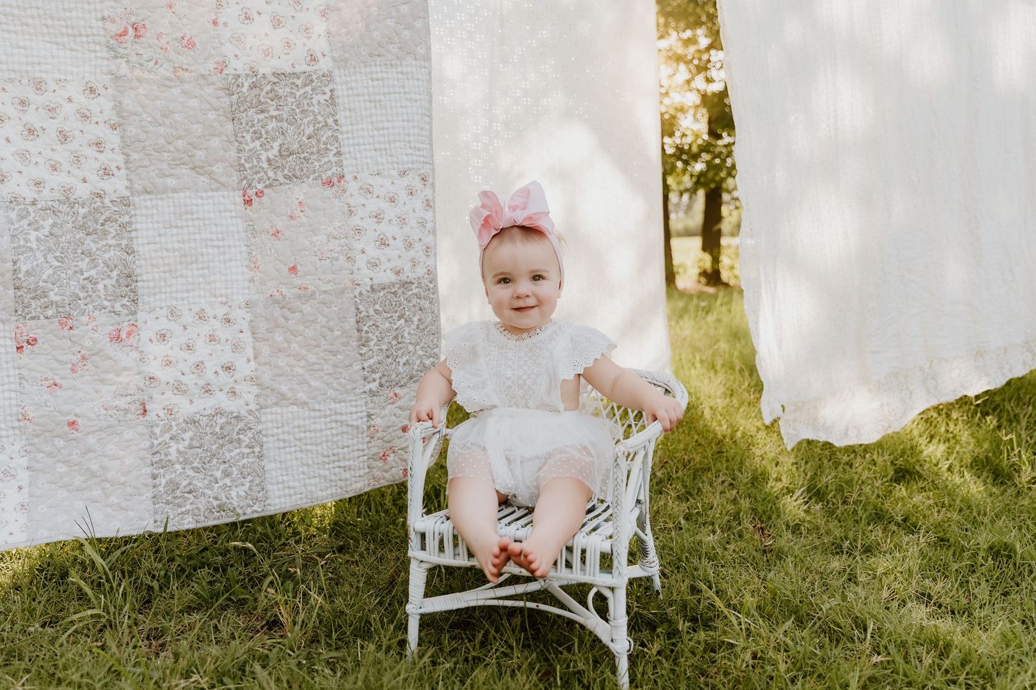 Baby girl in white dress and pink bow headband sitting on wicker chair outdoors with quilts hanging nearby.