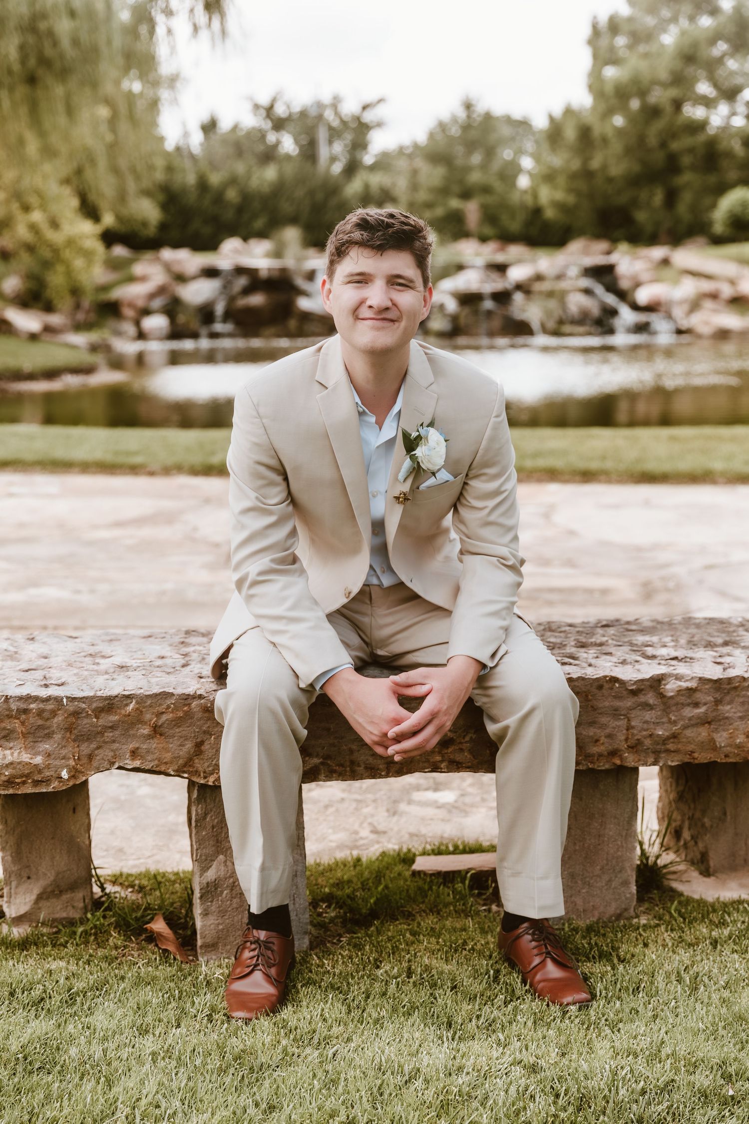 Young groom in beige suit with boutonniere sits on stone bench, smiling outdoors near a pond.