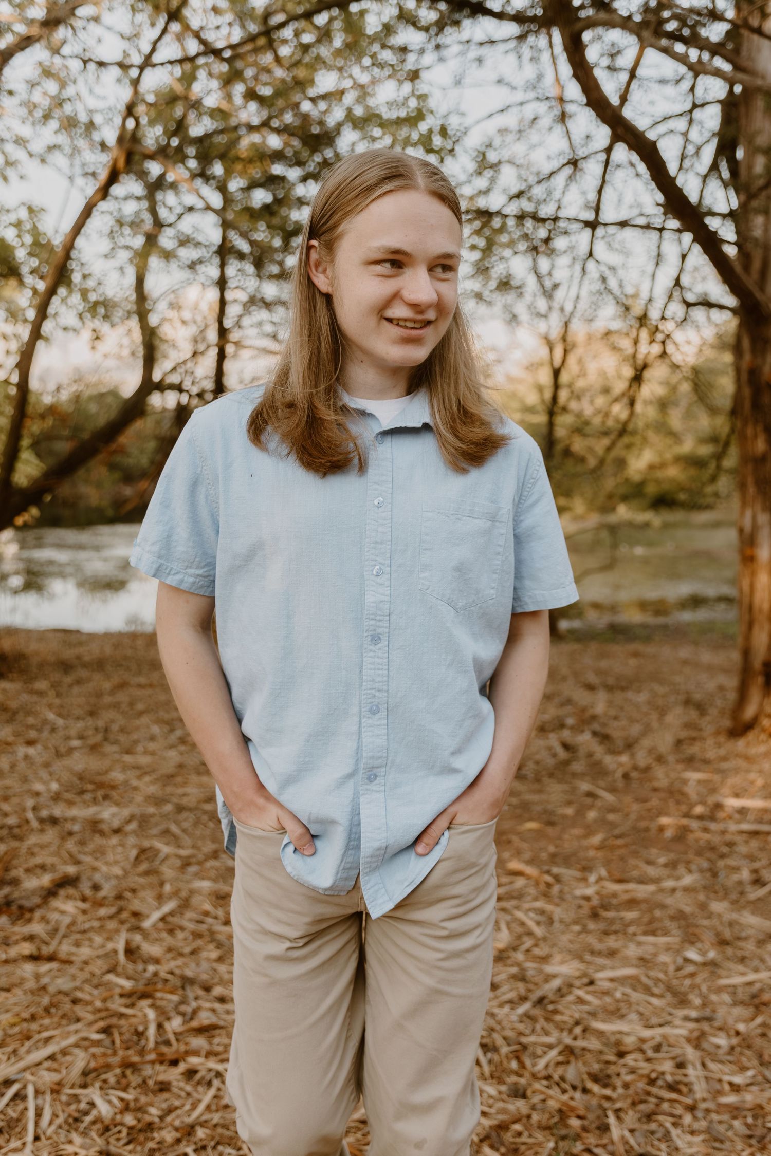 A person in a light blue button-down shirt and khaki pants stands outdoors in a natural setting during golden hour.