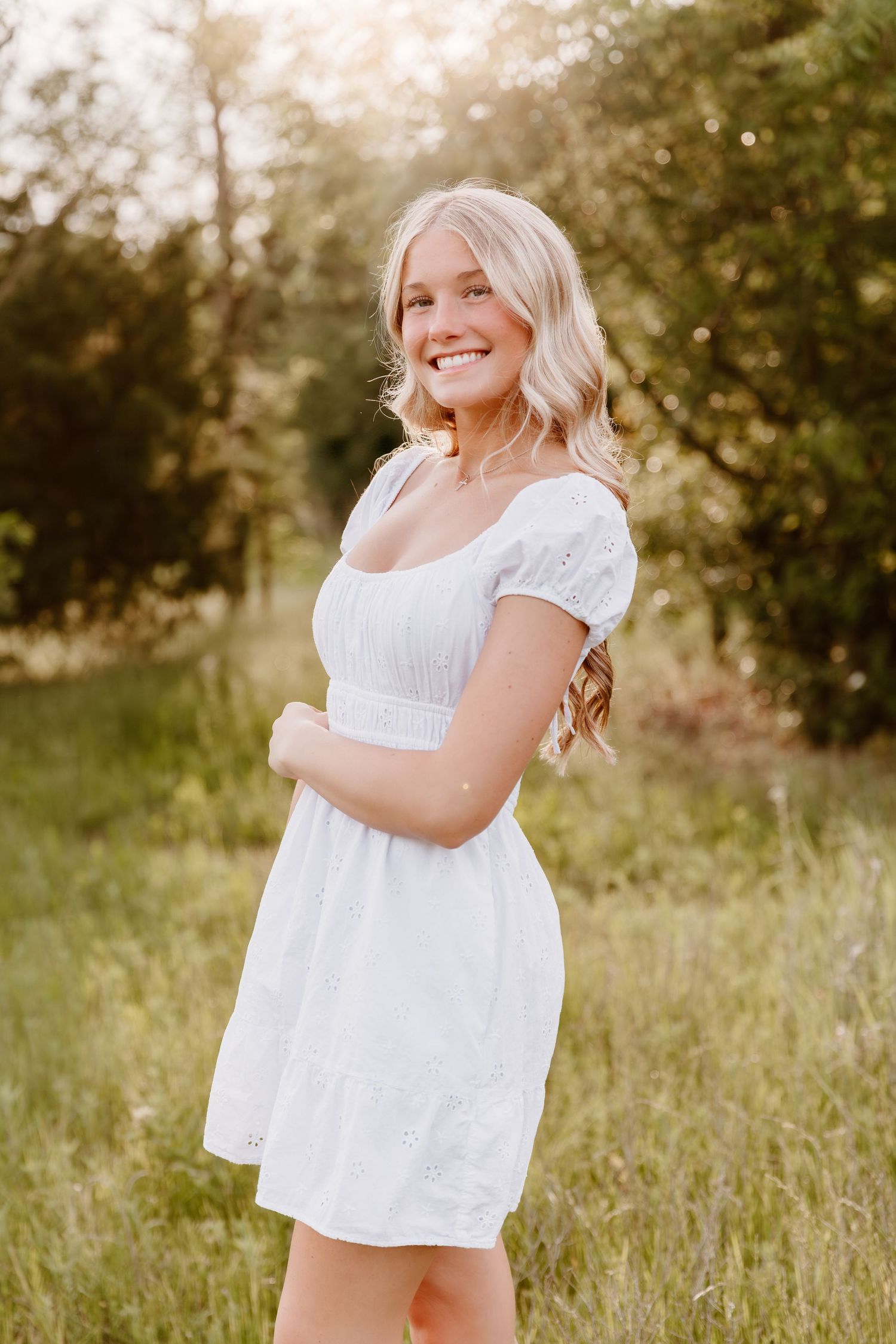 A person in a white summer dress stands joyfully in a sunlit meadow with tall grass and trees in the background.