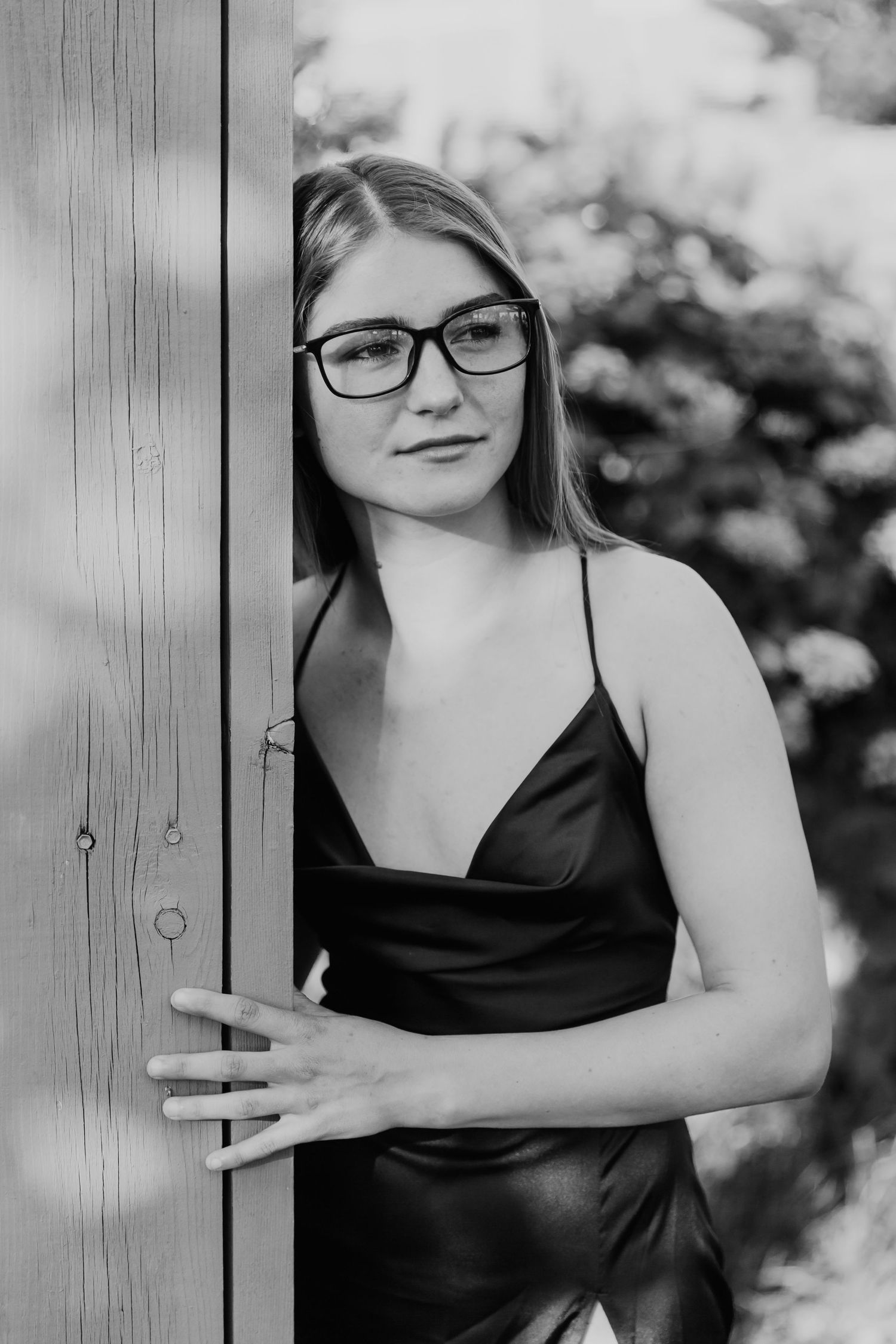 Woman in black dress strikes creative artistic pose while leaning against rustic wooden wall in black and white photo.