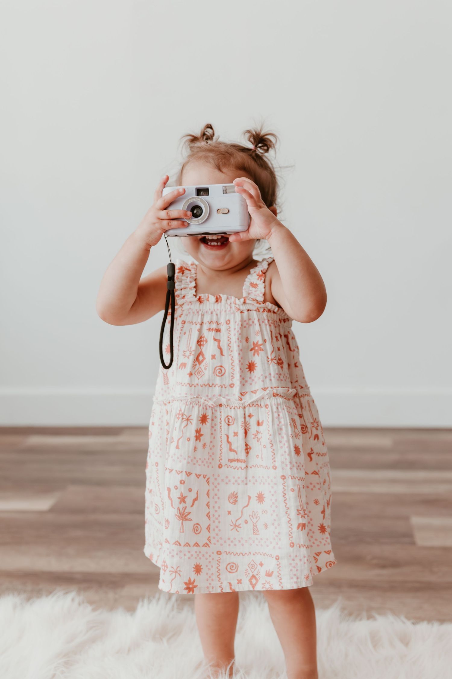 Small child in floral dress holding and playing with a vintage film camera against a light colored backdrop.
