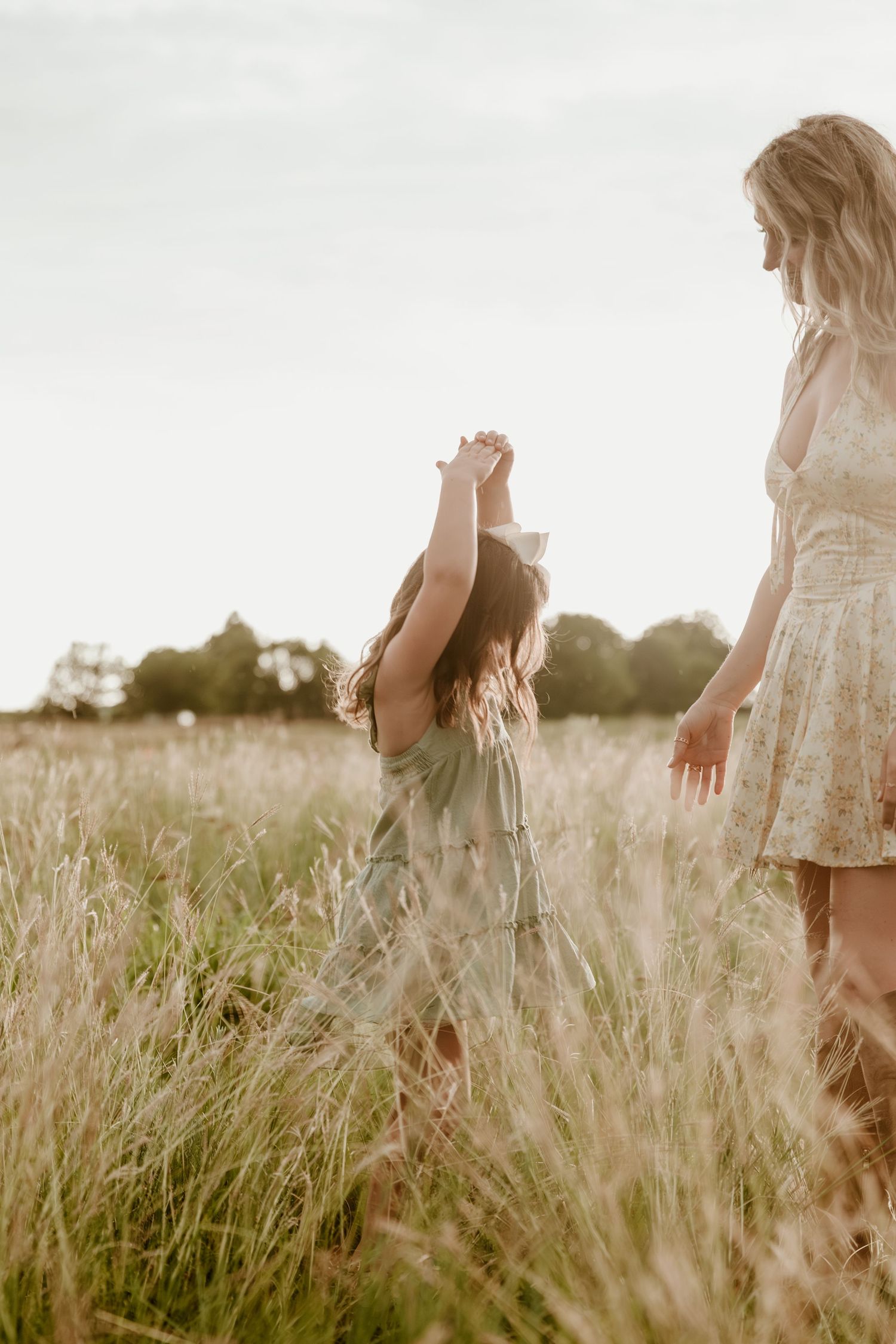 Sun illuminates tall seeded grass with figures in white lace dresses walking through a dreamy summer meadow.