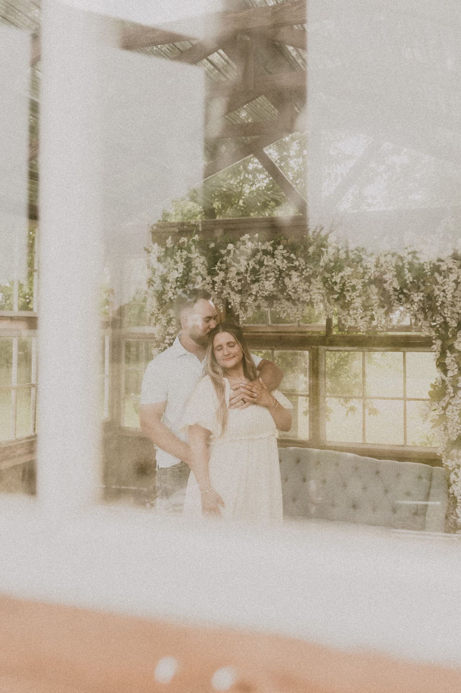 Romantic couple embracing in a sunlit greenhouse venue with floral decor and tufted sofa backdrop.
