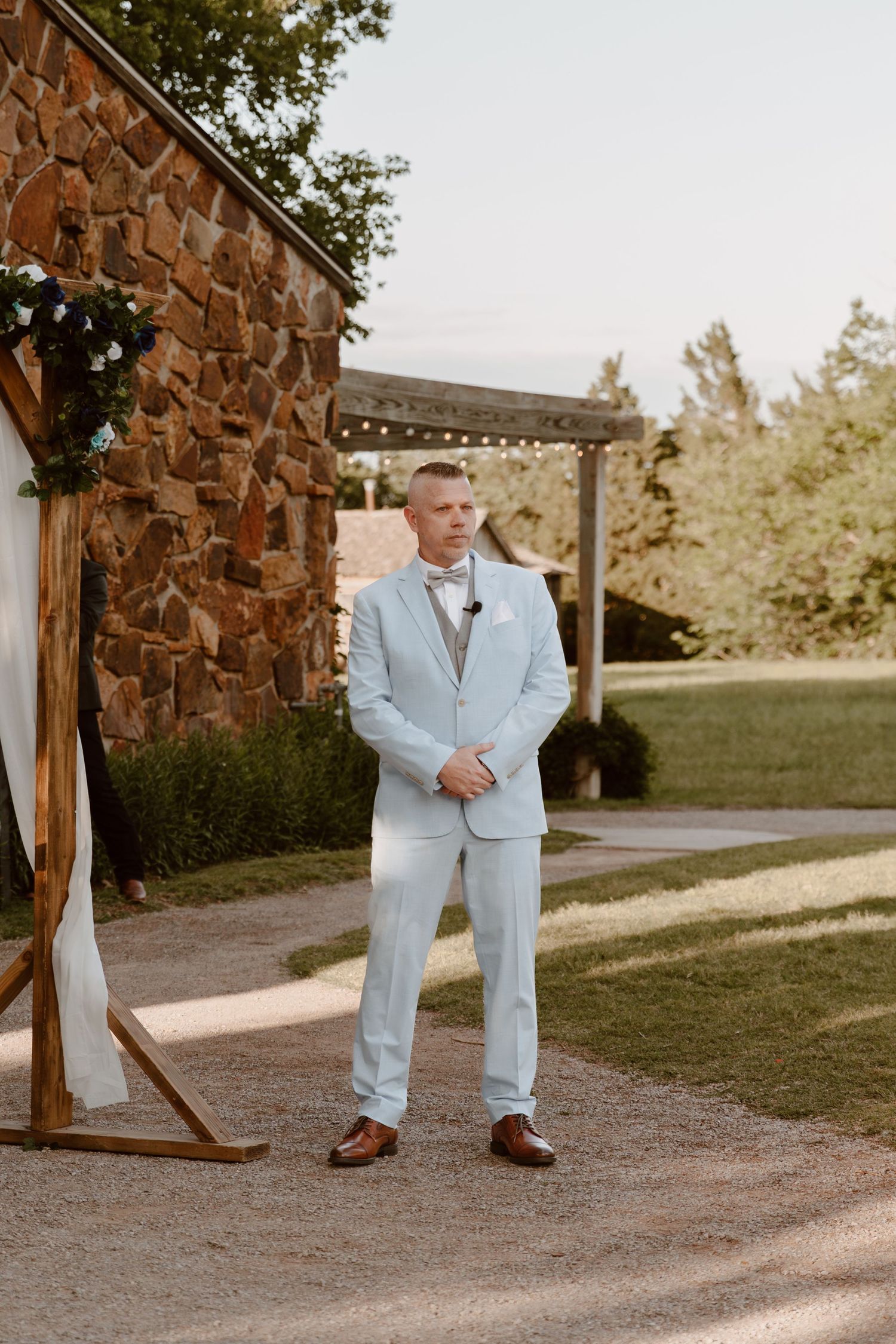 A groom in a light blue suit stands waiting at a rustic stone venue decorated with string lights.