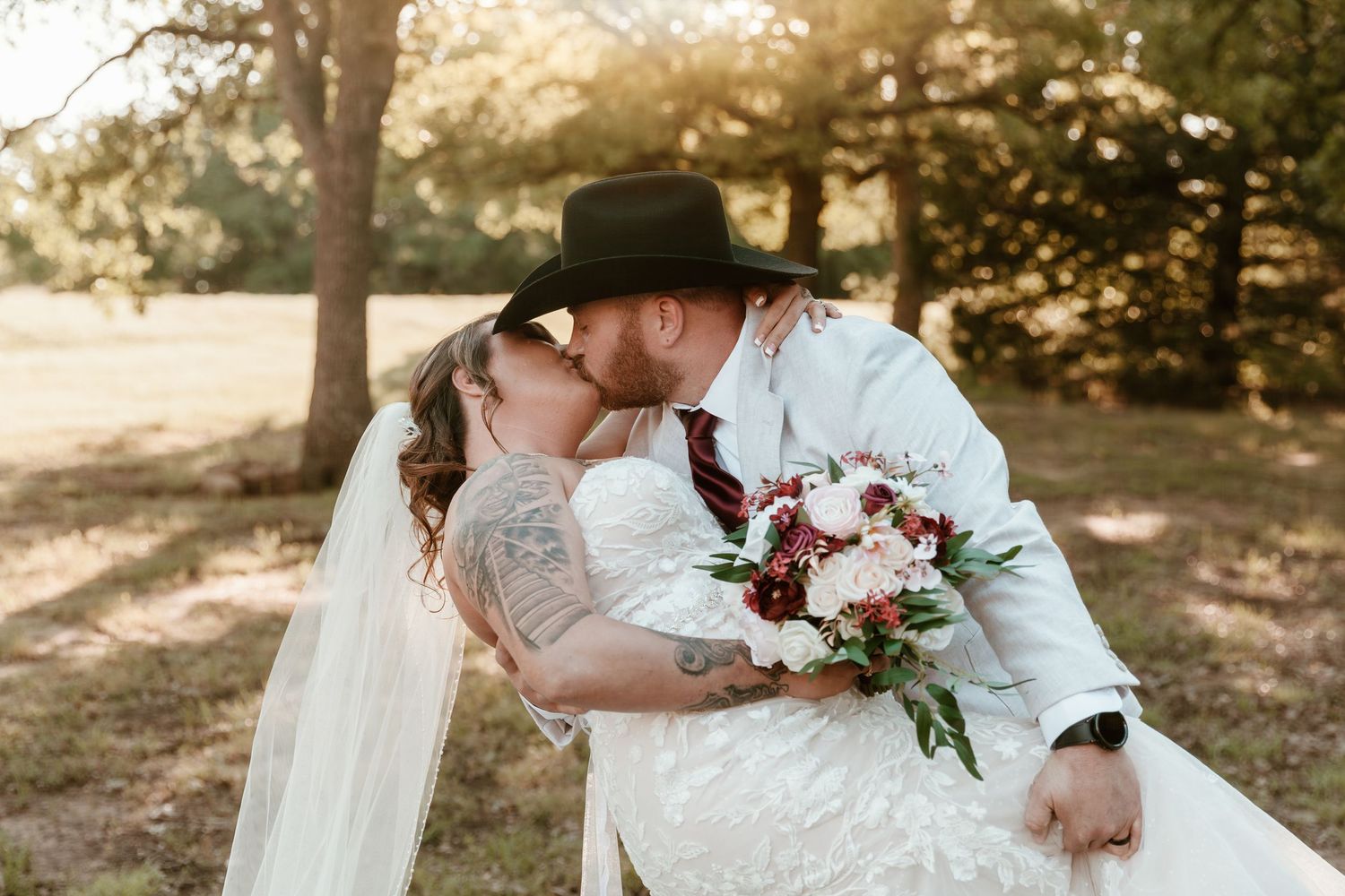 A romantic couple in western wedding attire share a kiss while holding a pink and white floral bouquet at sunset.