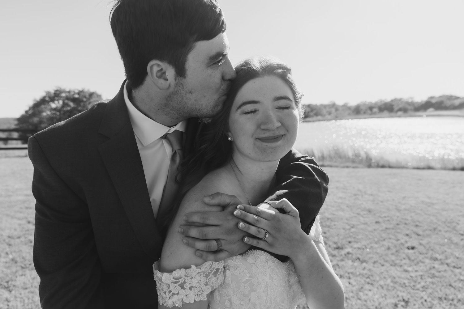 Black and white photo of a groom kissing bride on cheek while she smiles joyfully outdoors.