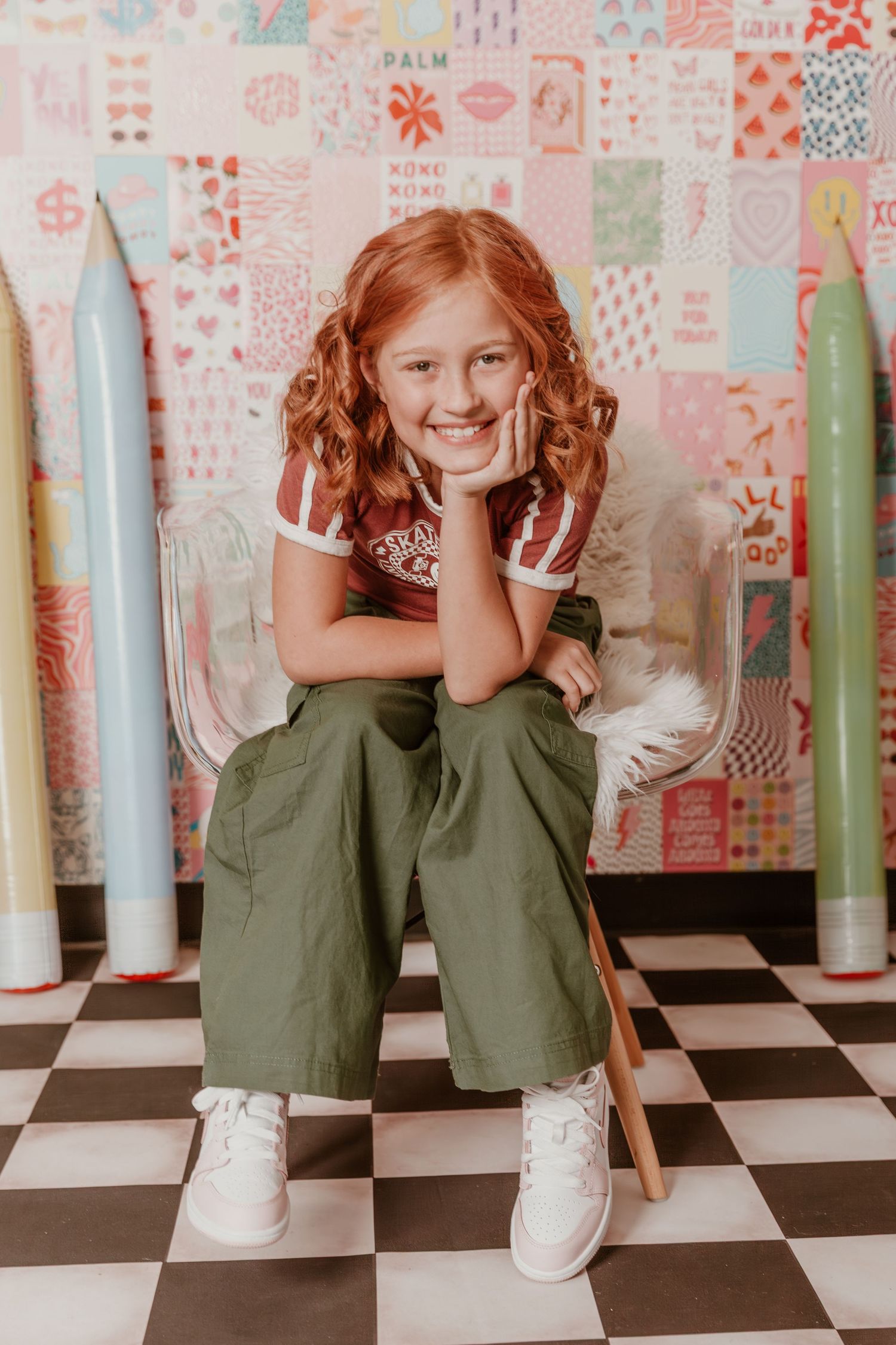 Candid portrait of someone sitting between giant decorative pencils against a whimsical patterned wall on checkered floor.