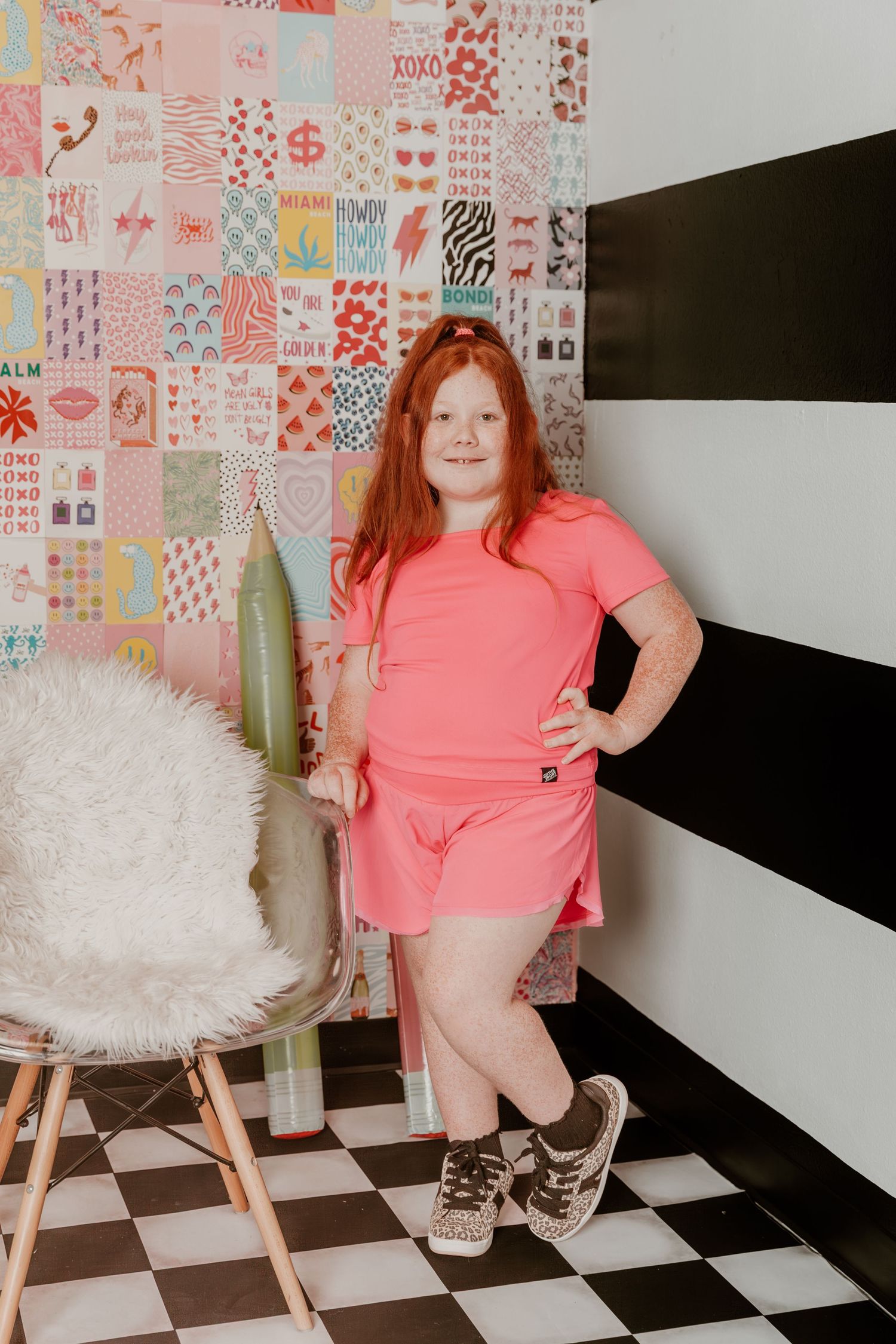 A pink-clad individual stands next to a furry white chair against a patterned wall and black and white striped backdrop.