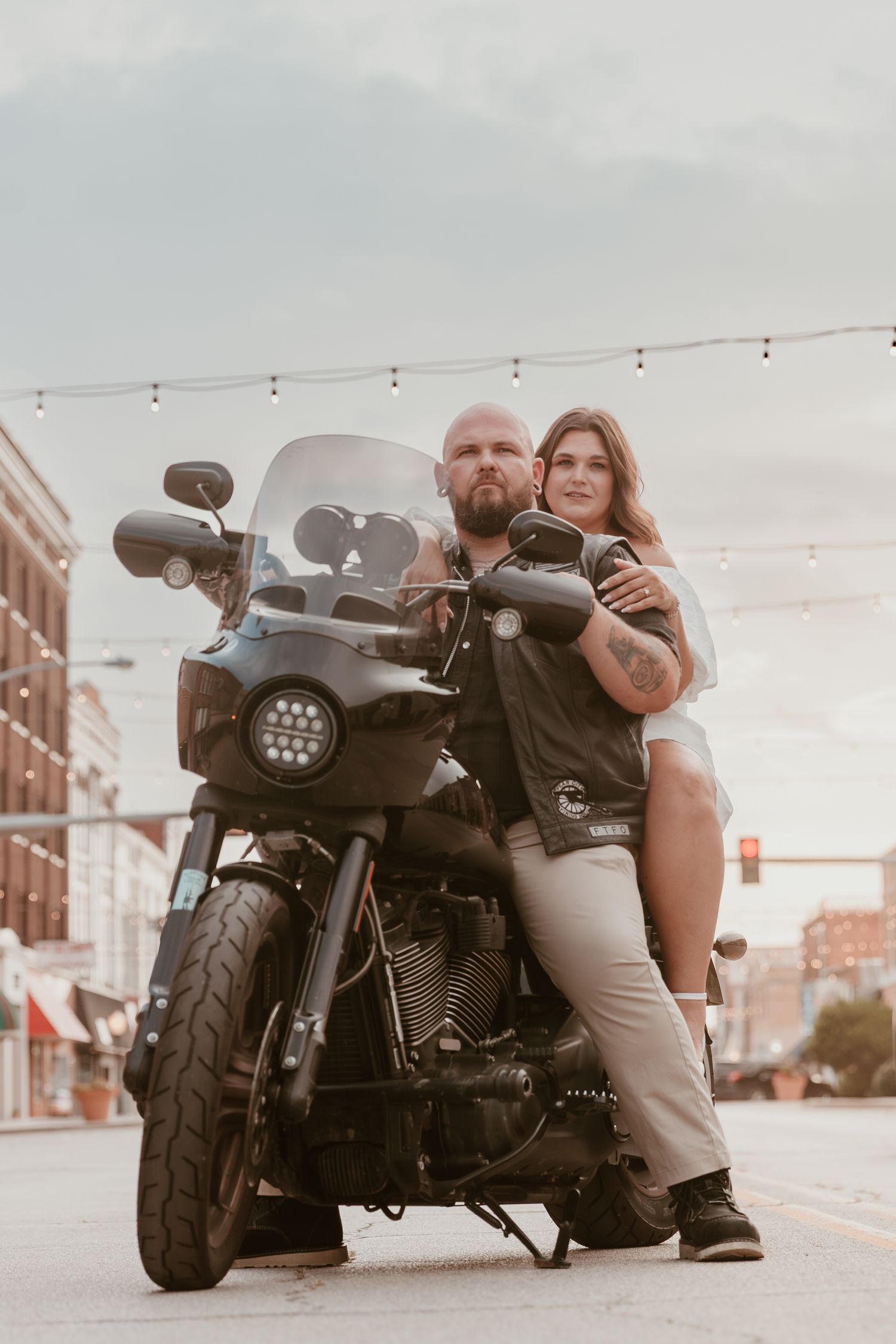 A couple poses on a motorcycle on a downtown street with string lights overhead and brick buildings in the background.