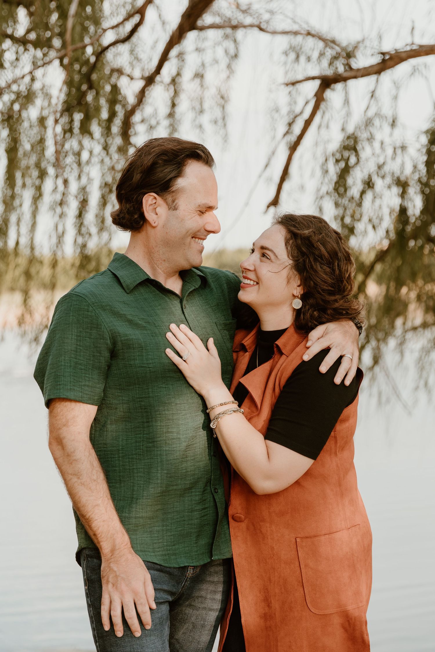 A couple shares a tender moment laughing together outdoors with willow trees in the background.