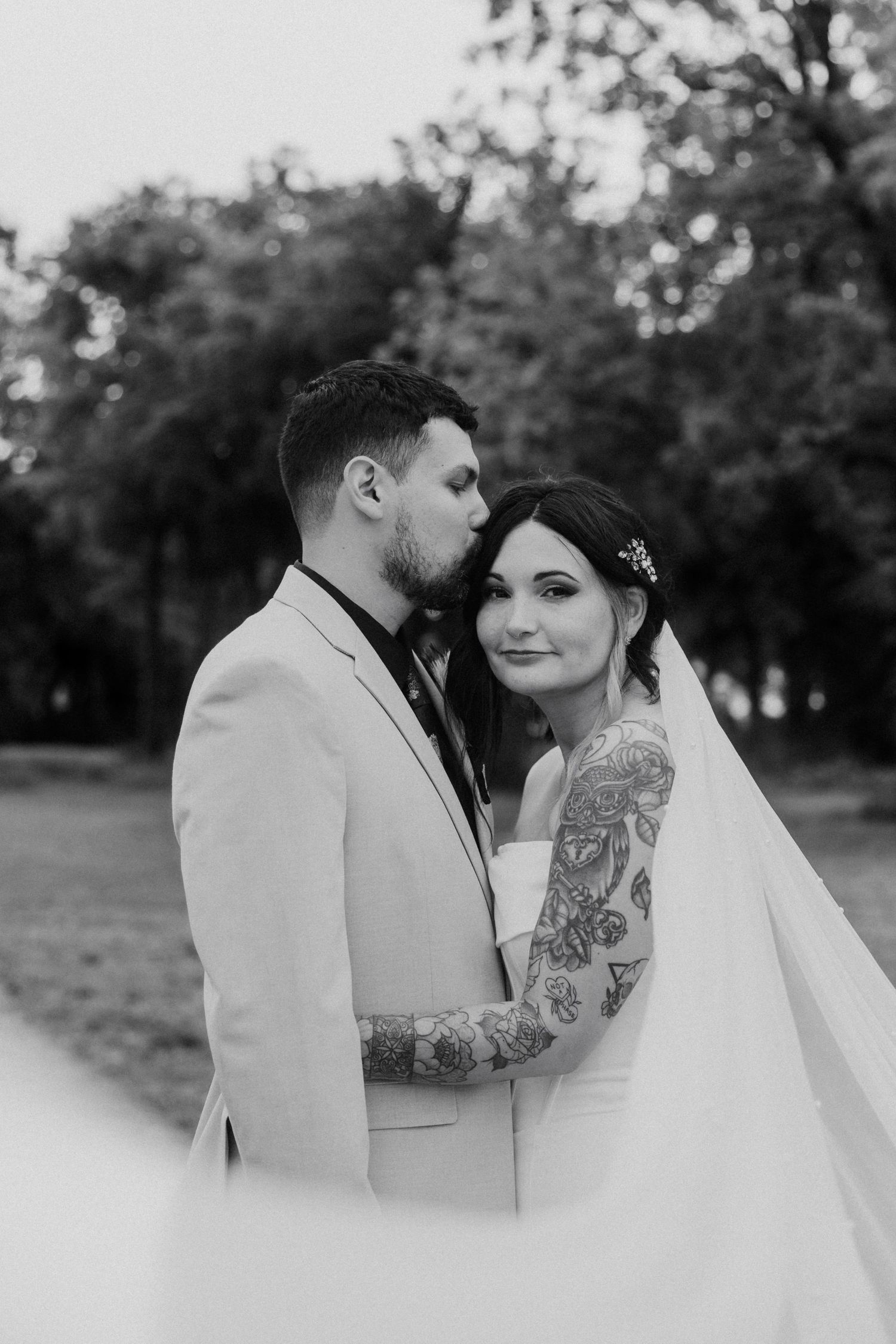 Black and white wedding photo of tattooed groom kissing bride on cheek outdoors among trees.