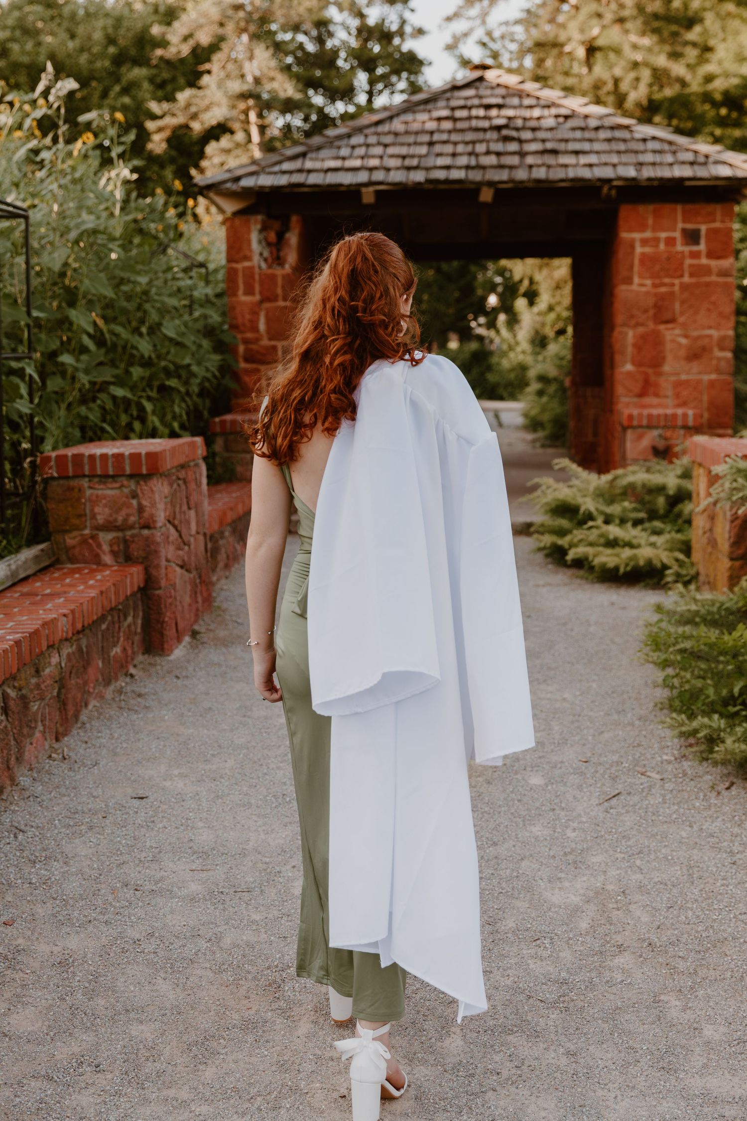 Walking figure in olive pants with flowing white garment on gravel path between brick garden structures.