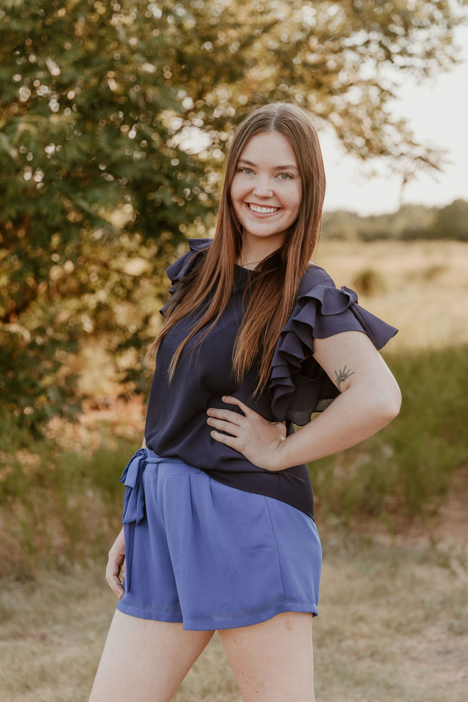 Outdoor portrait of a person in a navy ruffled top and blue shorts posing against a natural sunlit backdrop.