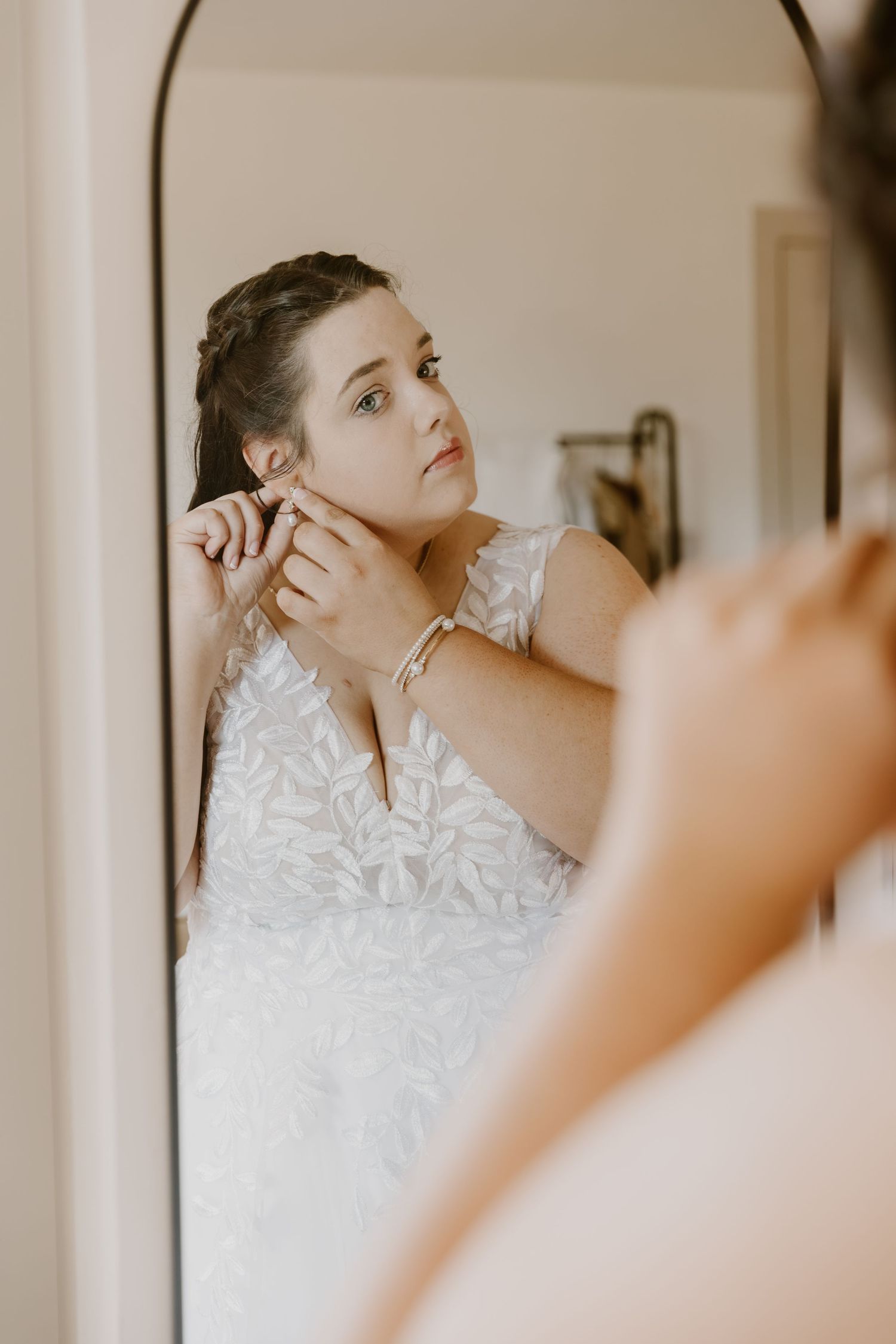 Bride putting on earrings while looking in mirror wearing white floral lace wedding dress.