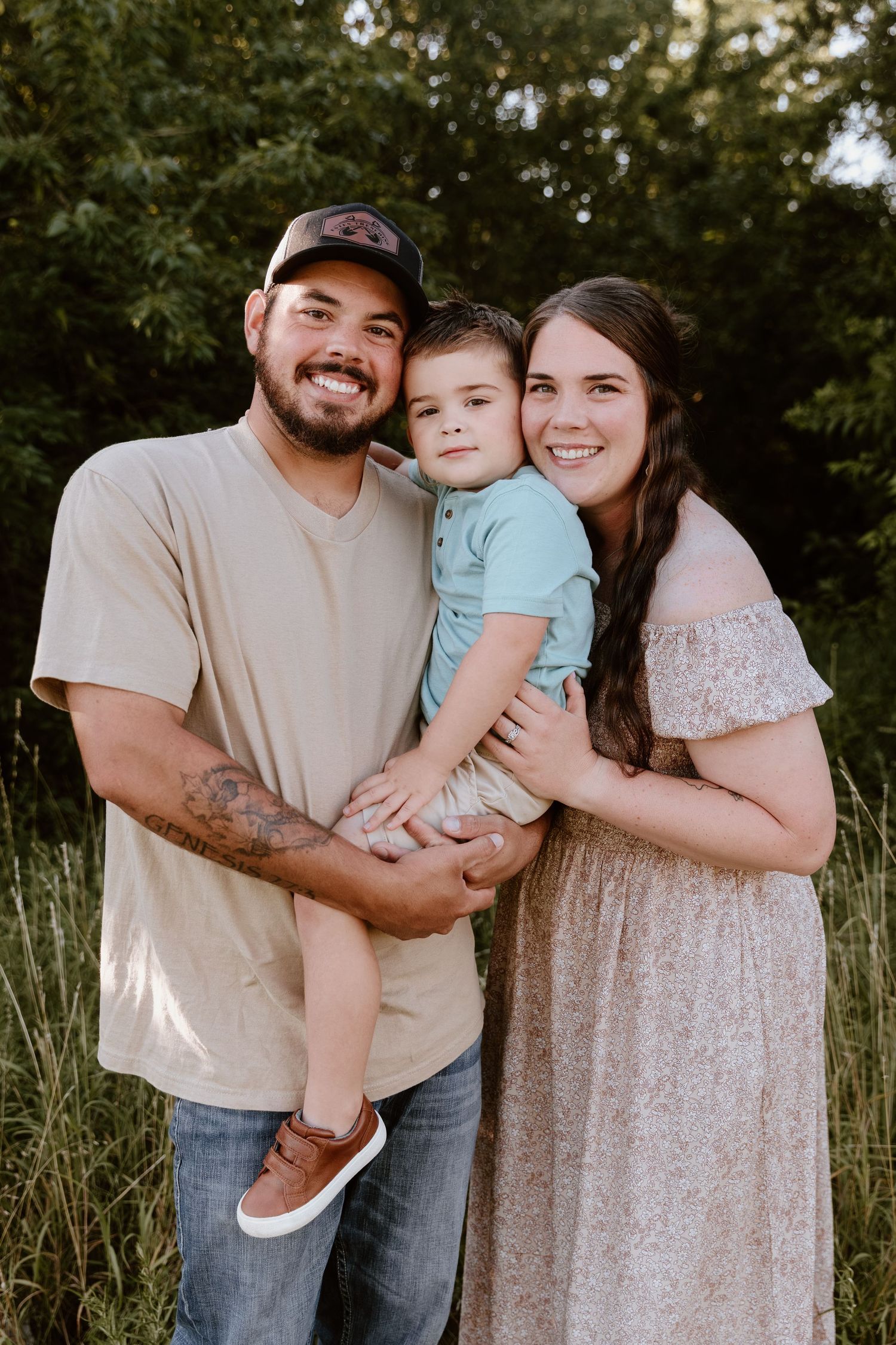 Happy young family with toddler boy posing outdoors in a grassy field with lush green trees in background.
