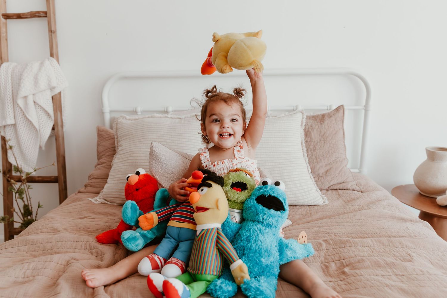 A child sitting on a bed surrounded by colorful plush toys including Cookie Monster and Elmo stuffed animals.