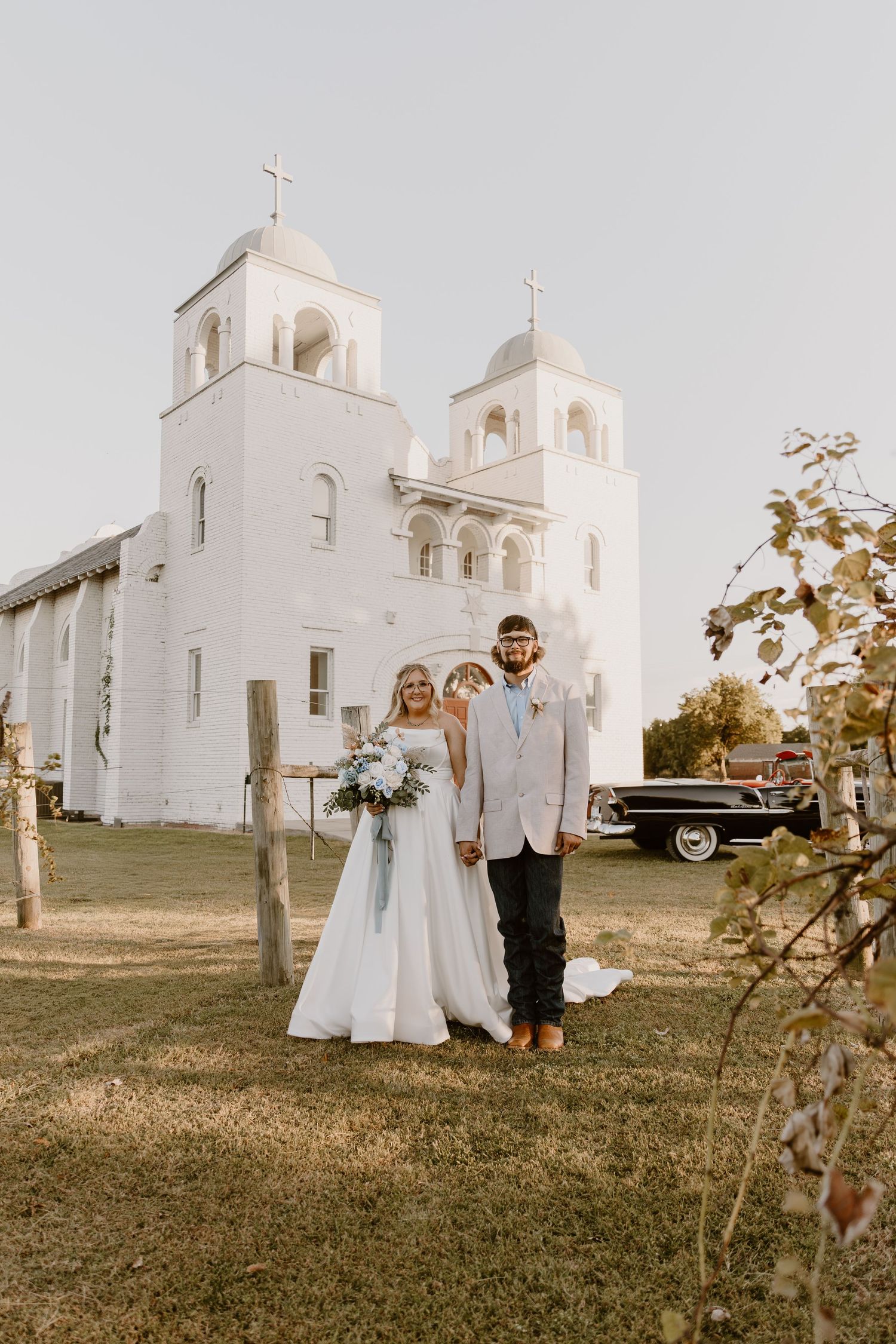 Wedding party poses in front of white Spanish colonial mission church on grassy hill at sunset.