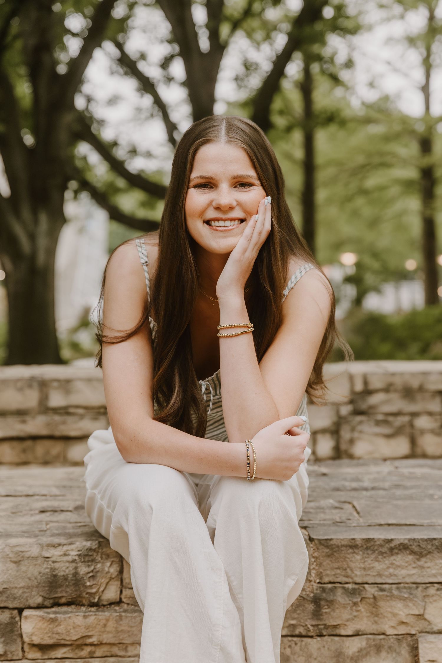 Smiling young woman with long brown hair sitting on stone steps outdoors, wearing white pants and gold bracelets.