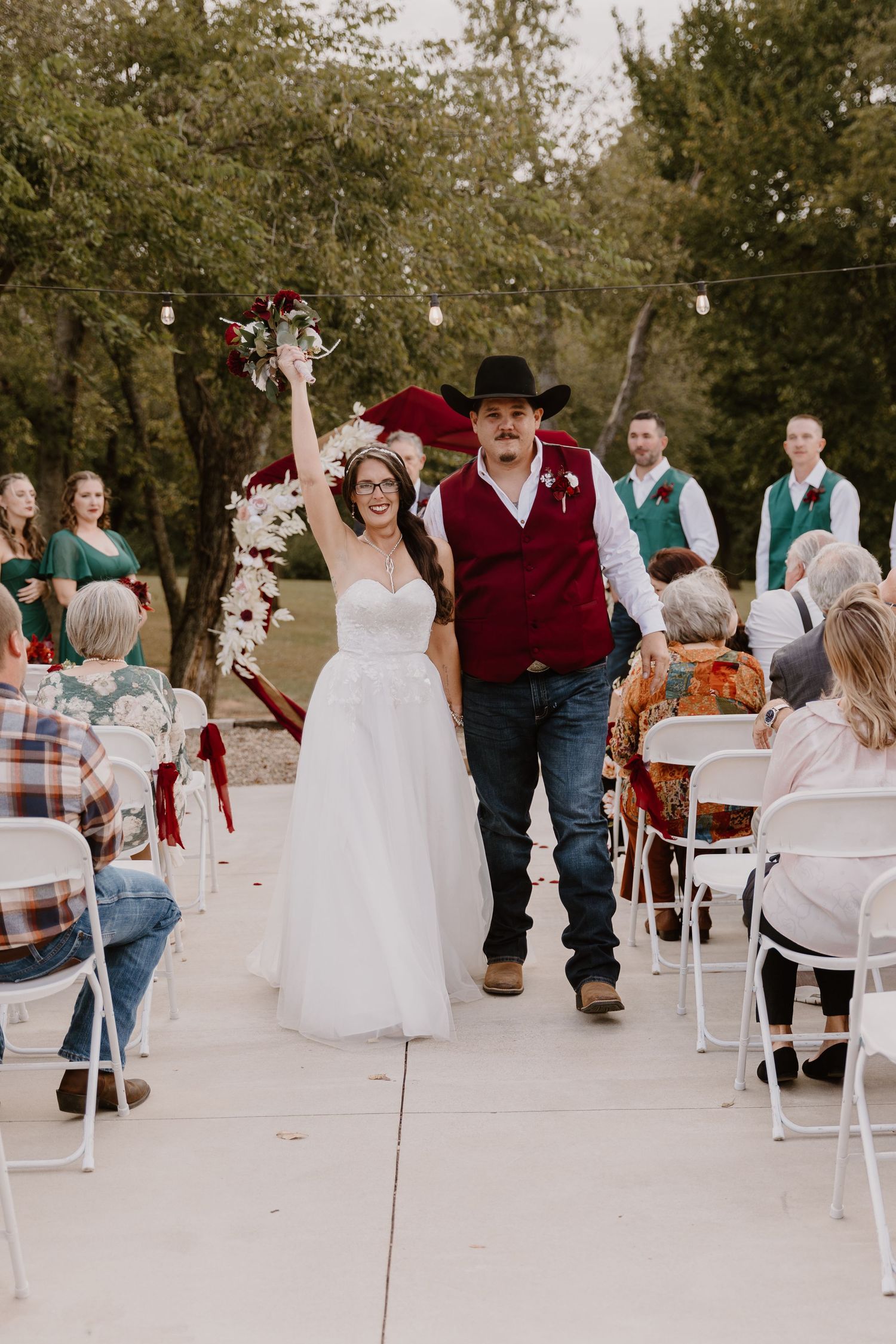 Outdoor wedding reception with guests seated at white chairs and tables under string lights.