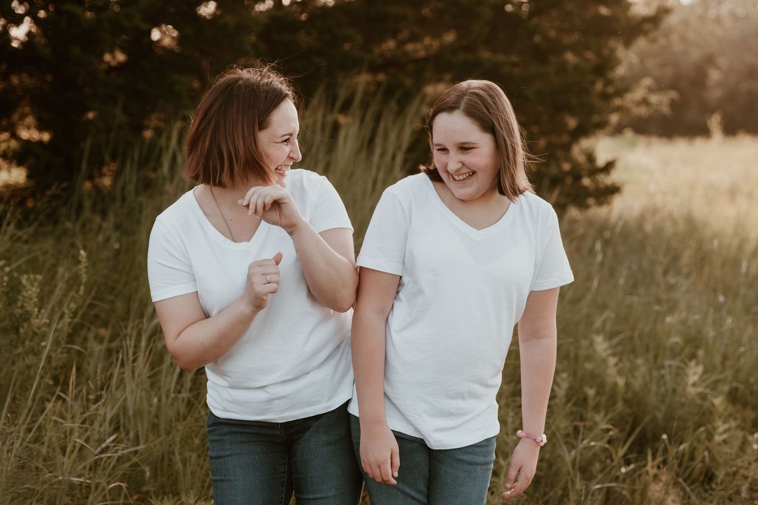 Two people in white t-shirts share a laugh while standing together in a sunlit field at golden hour.