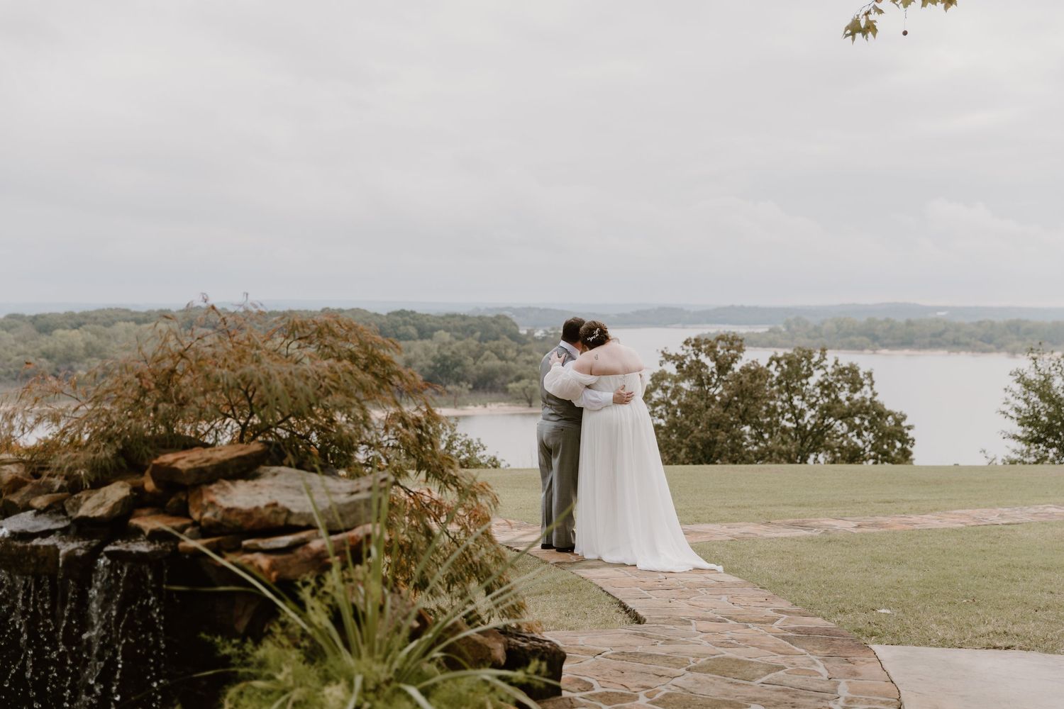 A bride and groom share a romantic embrace on a stone terrace overlooking a scenic lake on their wedding day.