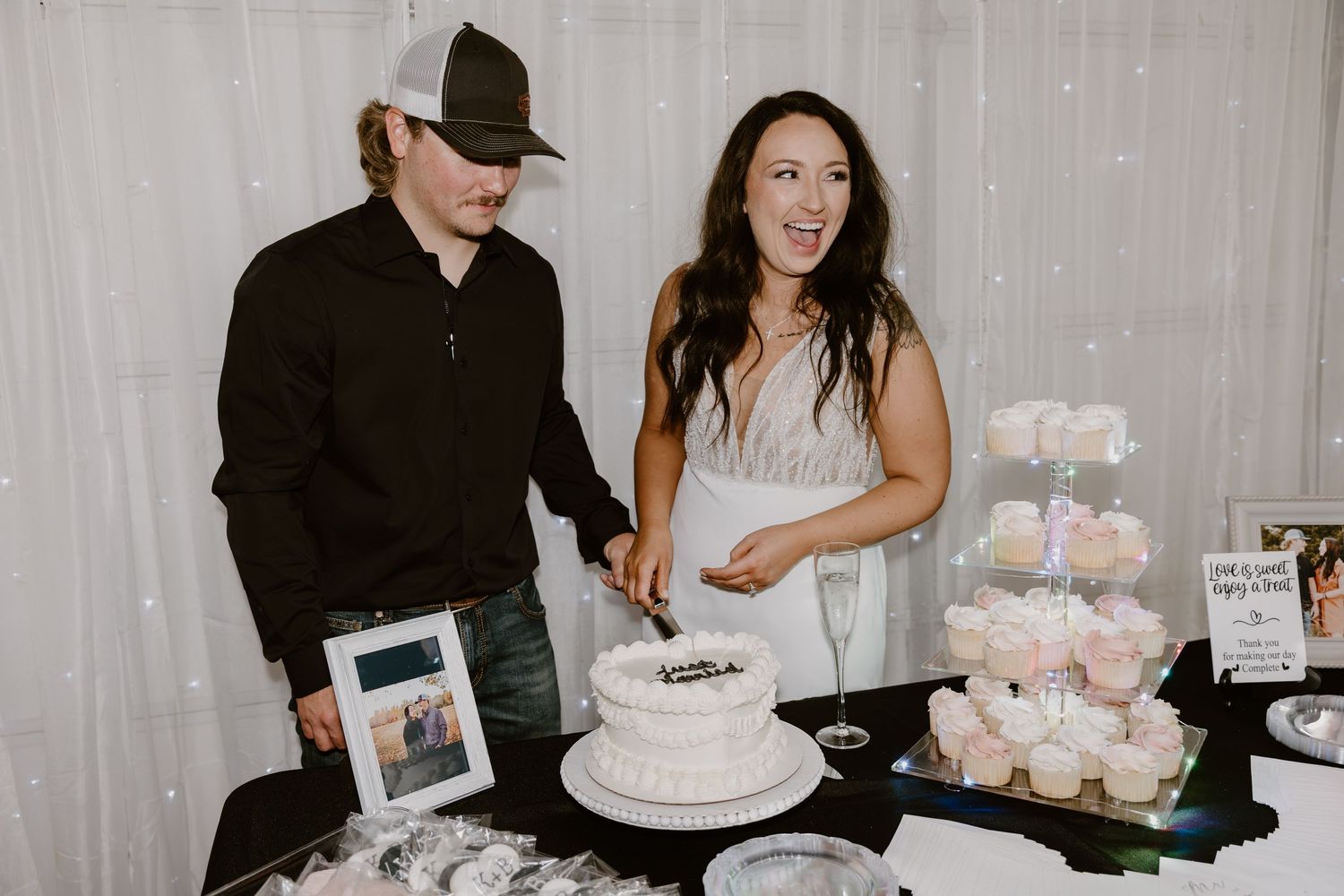 Wedding guests gather around a dessert table with cupcakes and treats at an indoor reception.