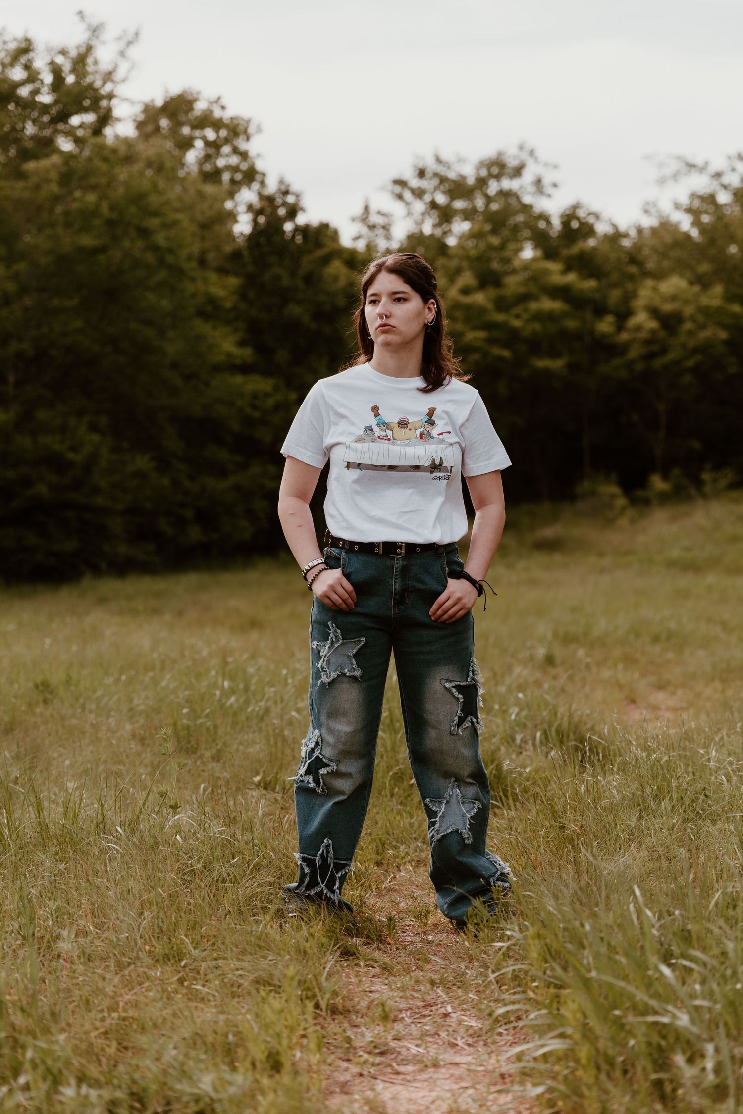 Person standing in grassy field wearing white t-shirt and distressed cargo jeans at sunset.
