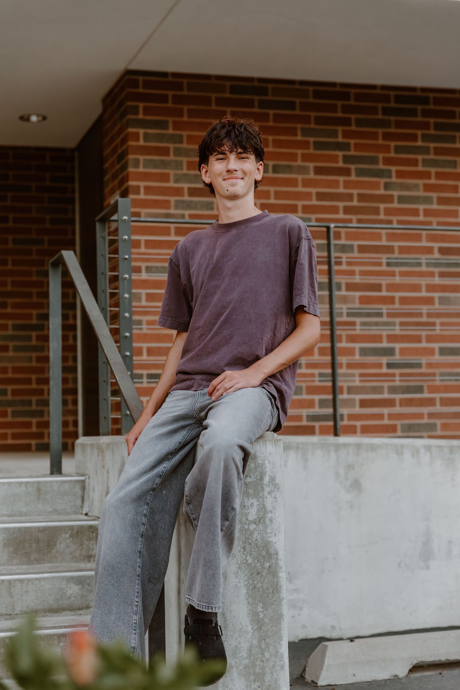 Person wearing a gray t-shirt sits on concrete ledge against brick wall in casual outdoor portrait.