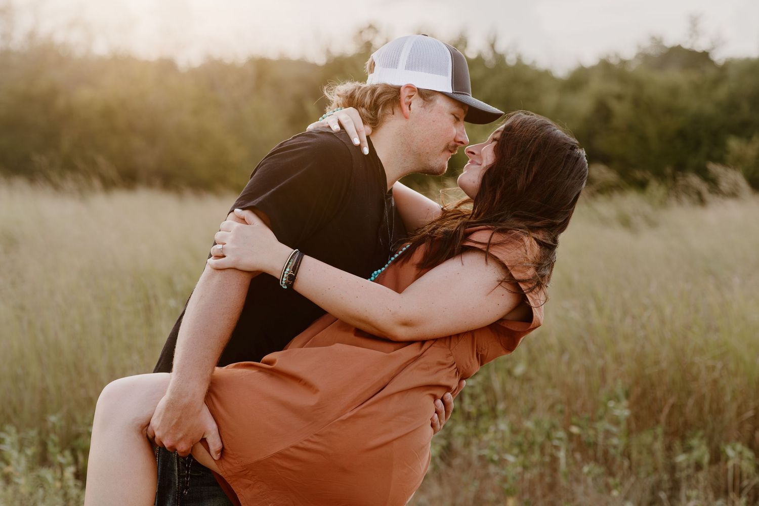 A couple embraces romantically in a sun-drenched field during golden hour while wearing earth-toned clothing.