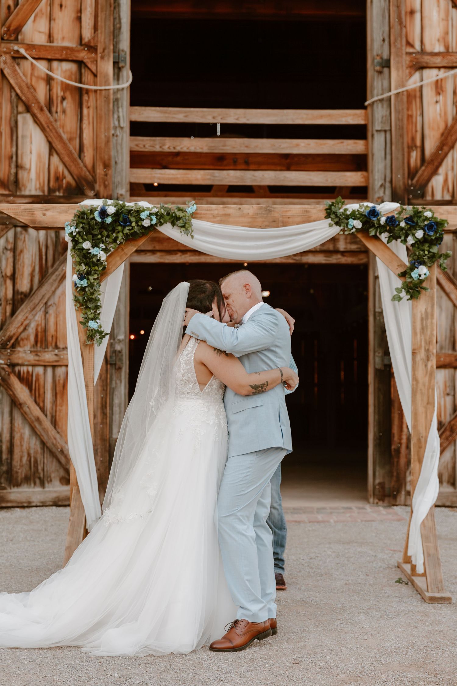 A romantic wedding scene at a rustic barn entrance decorated with white fabric draping and floral arrangements.