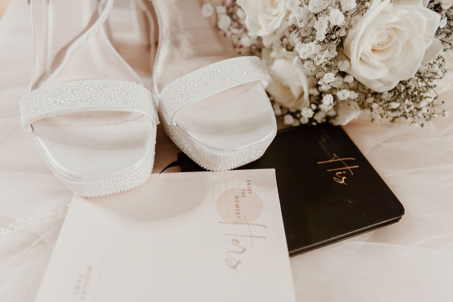 White wedding shoes and delicate rose bouquet arranged with invitation and passport in soft natural light.