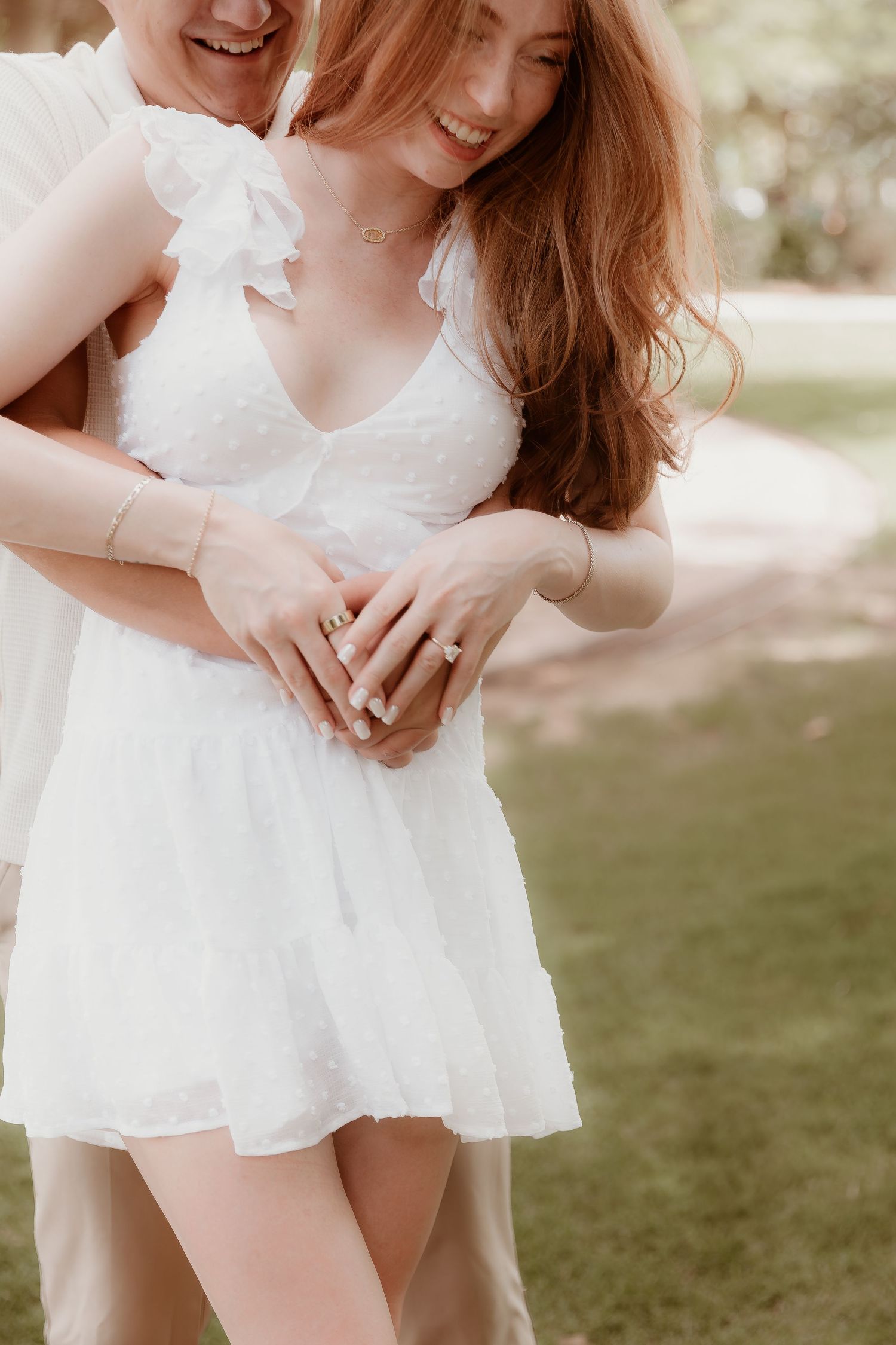 Couple embraces outdoors as sunlight filters through trees, with focus on white lace dress details and intertwined hands.