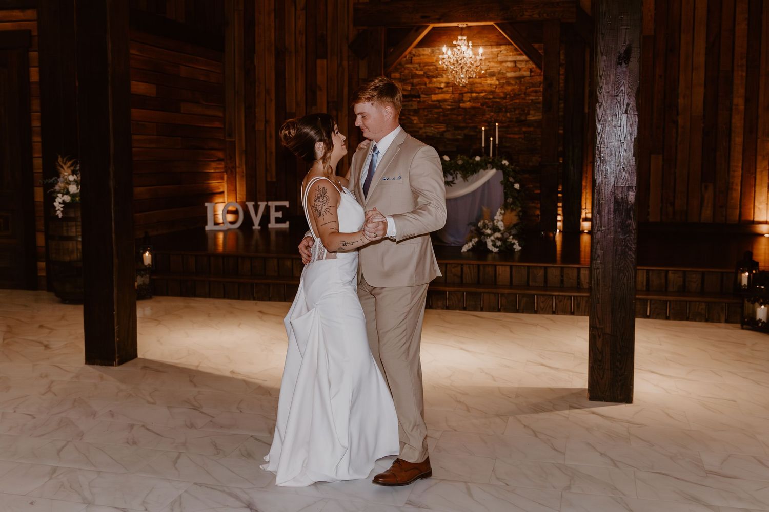 A couple shares their first dance in a rustic wooden barn venue lit by warm ambient lighting and candles.