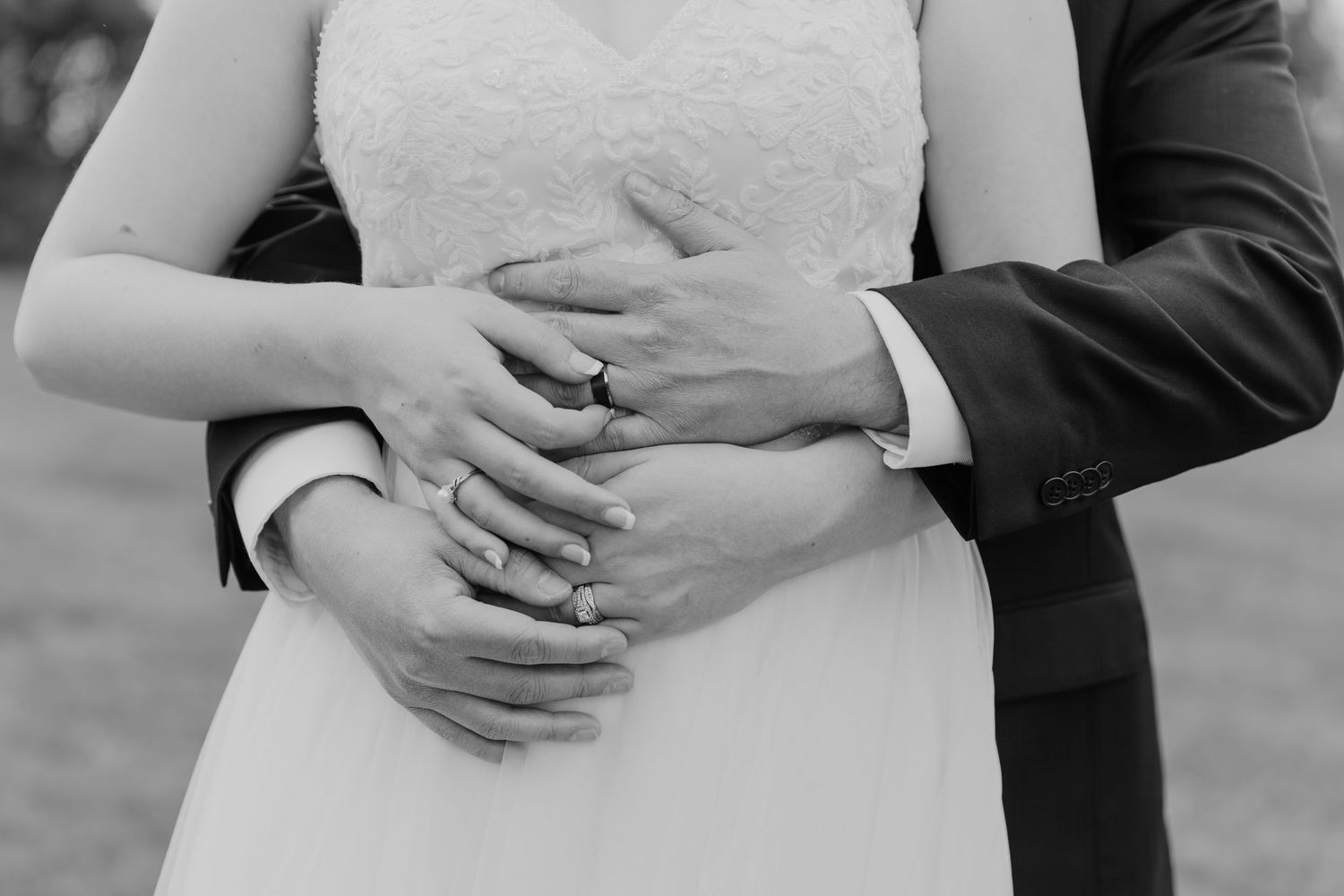 Black and white close-up photo of newlyweds hands embracing with wedding rings around a white dress.