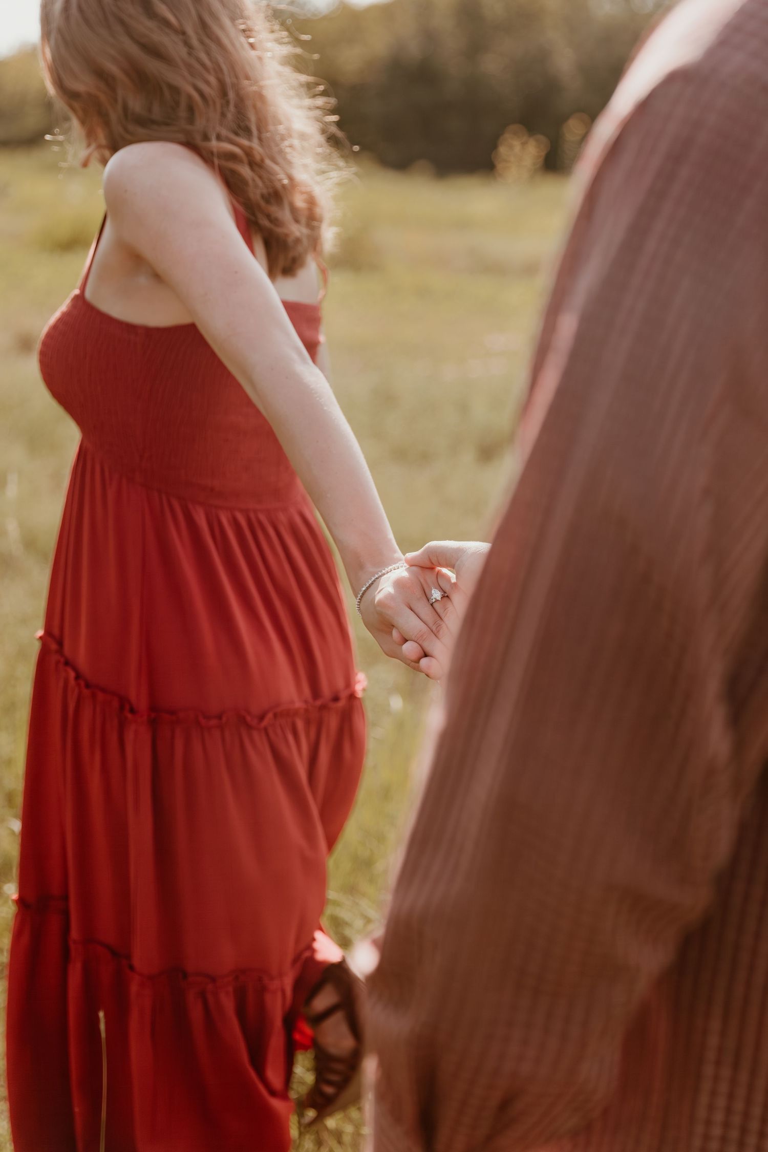 Close-up view of clasped hands against flowing red dress fabric in a sun-drenched meadow at golden hour.